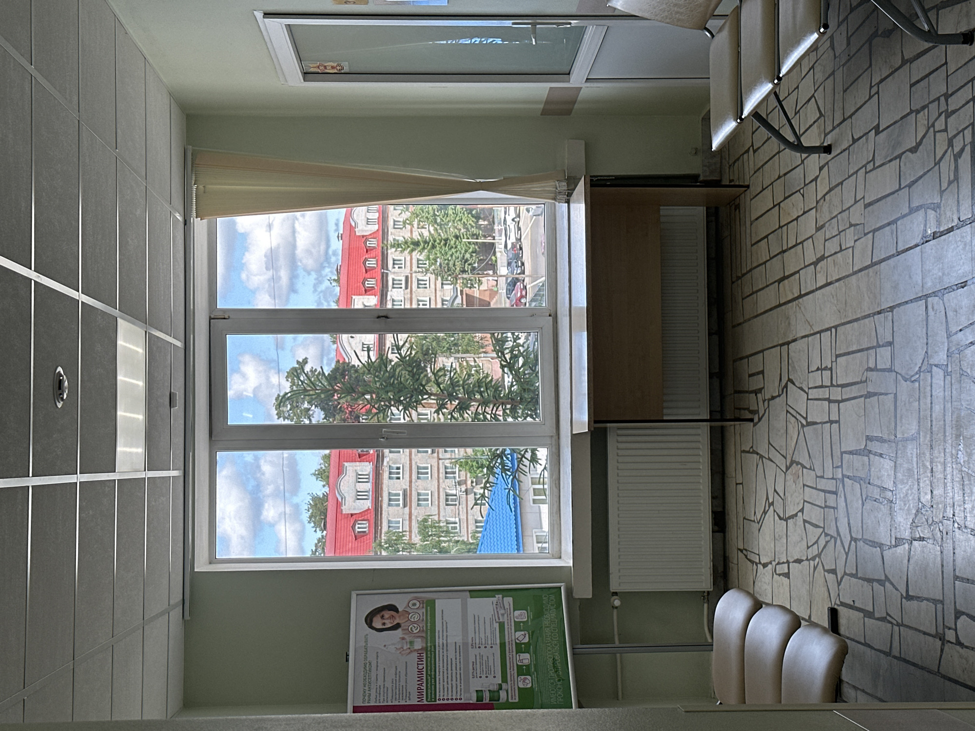 This image captures a brightly lit indoor waiting area, likely a medical office or public institution, located in Kazan, Russia. The room features light green walls and a ceiling composed of light gray square tiles with reflective silver strips. The floor and the wall on the right are covered with irregularly shaped, light-colored tiles, creating a mosaic-like pattern. Three light-colored, upholstered chairs with metal frames are visible against the right wall, suggesting a seating arrangement for visitors.
A large window dominates the left side of the room, offering a view of the outside. A partially drawn light beige curtain or blind is visible at the top of the window. Through the window, several multi-story buildings with reddish/orange pitched roofs and light-colored facades can be seen, interspersed with green trees. A street with parked cars is visible in the distance, and a prominent bright blue roof is present on a lower building. The sky is bright blue with scattered white clouds, indicating daytime.
Beneath the window, a long, light-colored wooden desk or shelf is present, with a white radiator mounted below it. On the wall beneath the window and to the left of the radiator, there is a framed, vertical poster. The poster displays a smiling woman, medical symbols, and images of product bottles. The most prominent visible text on the poster is in Cyrillic, reading "МИРАМИСТИН" (Miramistin), which is a known antiseptic drug. Other Cyrillic text, likely informational, is also present. The overall impression is one of a clean, functional public space during clear daytime weather.