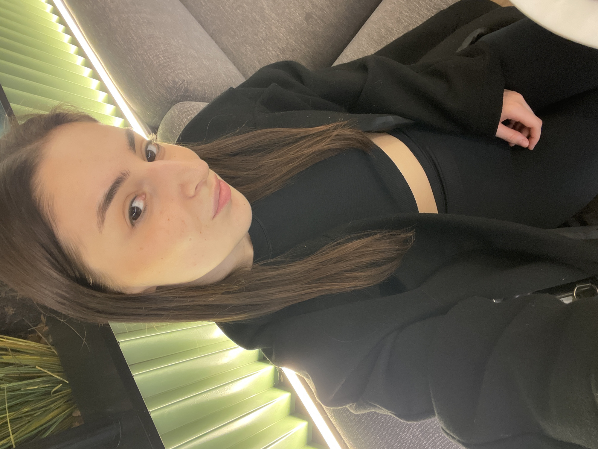 A young woman with brown hair is lying on a gray sofa indoors, wearing a black top and a black jacket. She has light skin with freckles. A wall with a green, linear design and bright lighting is visible behind her. The location is undetermined and not discernible as Moscow, Russia.