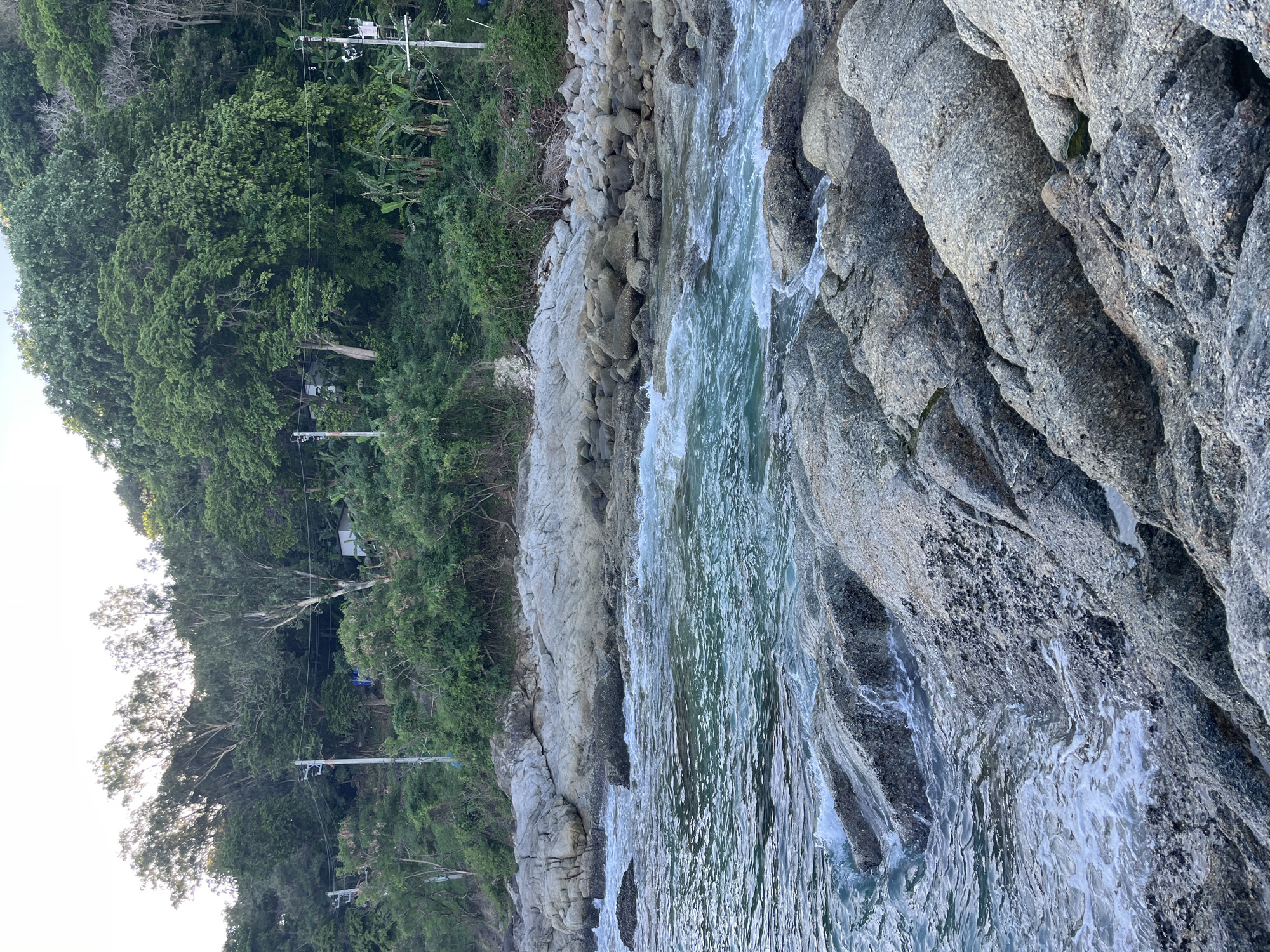 The image shows a rocky coastline with the turquoise water of the Andaman Sea in Rawai, Thailand. The foreground is dominated by large, grey rocks with rough textures, some partially submerged in the water. Waves gently crash against the rocks, creating white foam. In the midground, the water is clear with visible ripples and currents. The background features a steep, lush green hillside covered in dense vegetation, including trees and banana plants. Several utility poles with wires are visible on the hillside, indicating human presence. The sky is overcast with a soft, diffused light. No people are visible in the scene. The overall impression is one of a natural, tropical coastal environment.