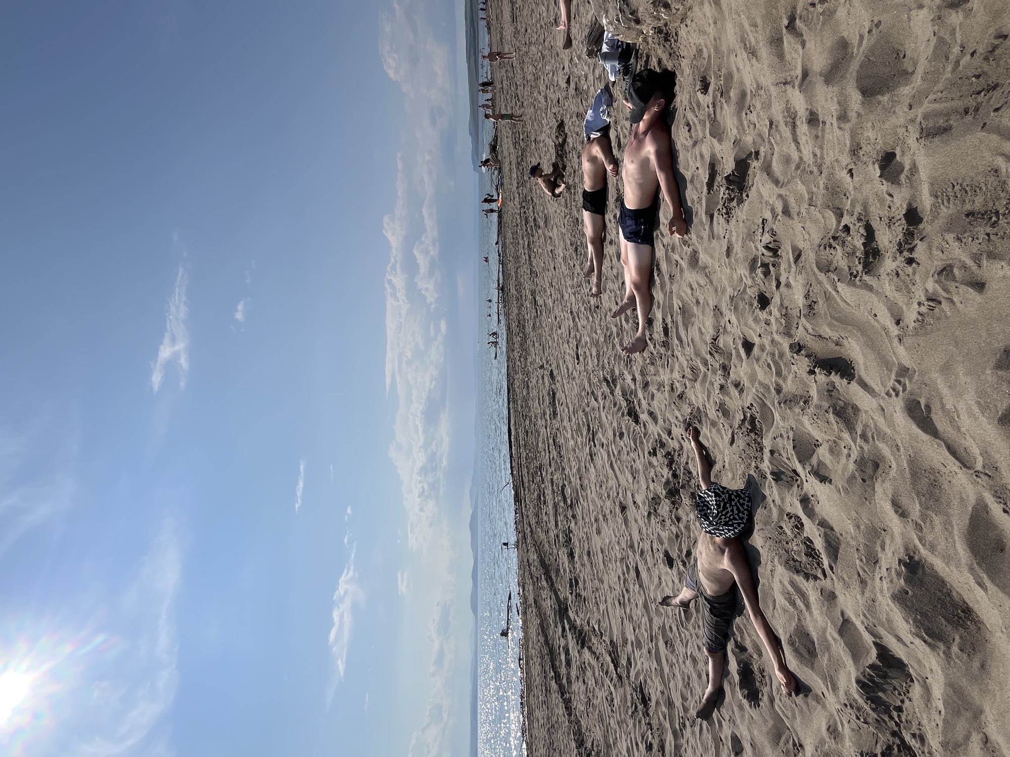 This media file depicts a sunny day at a beach, likely in Krasnoturansk, Russia, given the context. Several individuals are visible on the sandy shore. In the foreground, a person is lying on their back, wearing dark shorts and a patterned bandana. To their left, another person is also lying down, wearing dark swim trunks and a dark cap. Further in the background, more people are scattered across the beach, some appearing to be standing or walking. The sea stretches out beyond the beach, with distant hills visible on the horizon. The sky is blue with scattered white clouds. The sun is bright, casting shadows on the sand and creating a shimmering effect on the water, suggesting it is midday. The overall scene conveys a relaxed atmosphere of a typical summer day at the beach.