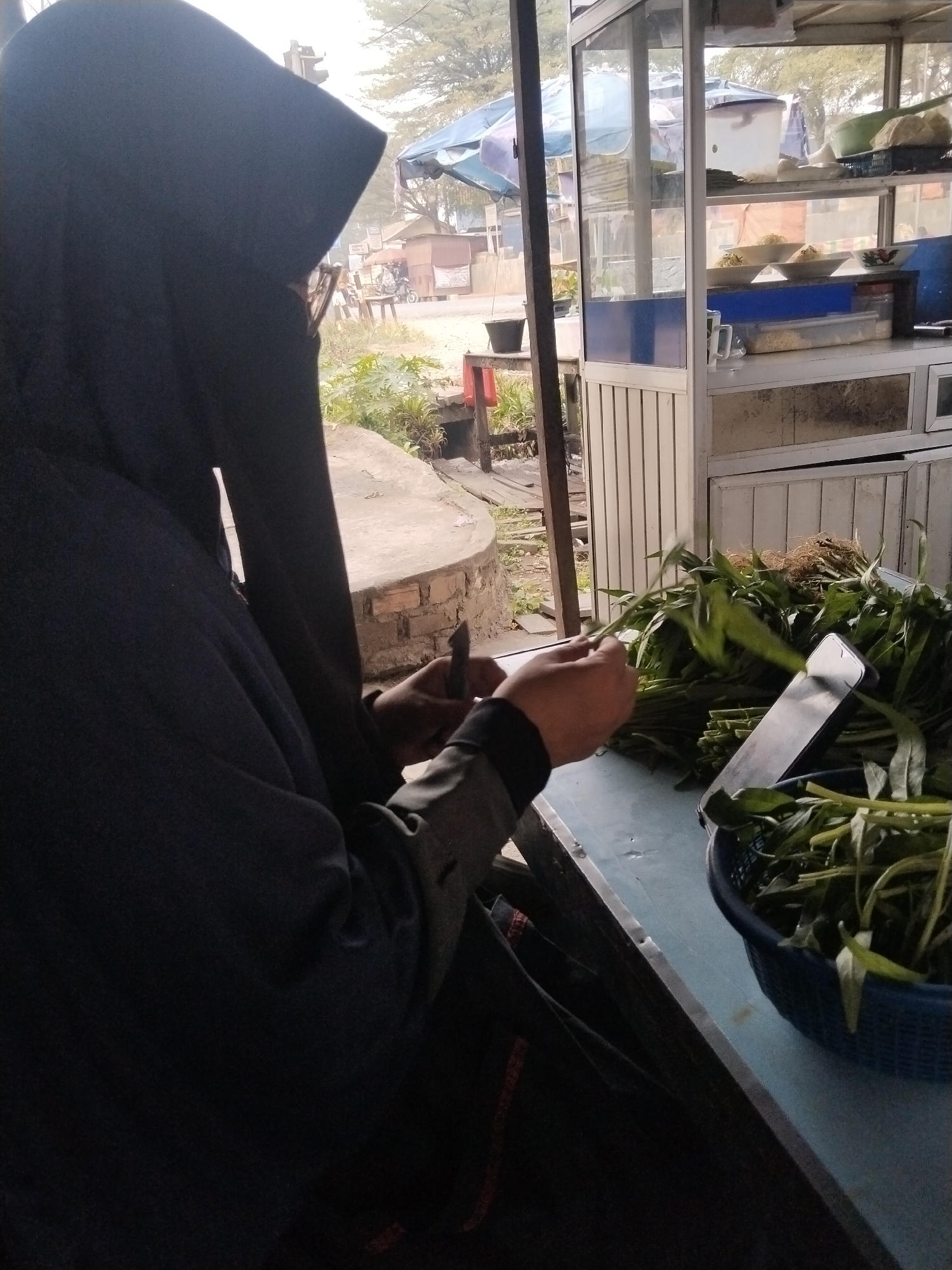 In Suka Ramai, Indonesia, a person wearing a dark hijab and niqab is seated at a blue table. Their hands are visible as they hold a knife and prepare a large bunch of leafy green vegetables, likely water spinach, which are in a blue basket to the right. A smartphone is propped up next to the vegetables. Behind the table is a food stall with shelves displaying food items in bowls and containers, and a cooler. The background suggests an outdoor setting with a street, trees, and a few buildings. The overall scene depicts food preparation at a roadside stall.