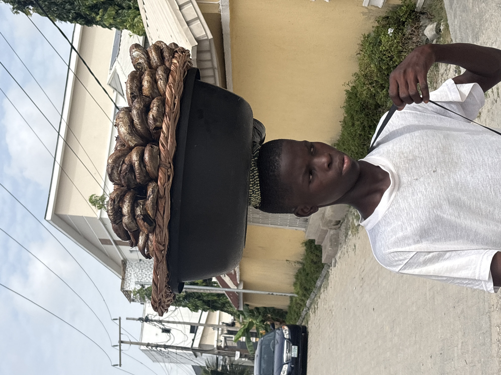 A young man walks along a paved street in Igboefon, Nigeria, carrying a large, round basket filled with smoked fish on his head. The basket is black with a brown woven rim. He wears a white T-shirt and has a patterned fabric head wrap beneath the basket. Power lines and buildings are visible in the background, along with a parked blue car. Green shrubbery and trees line the street. The sky is overcast.