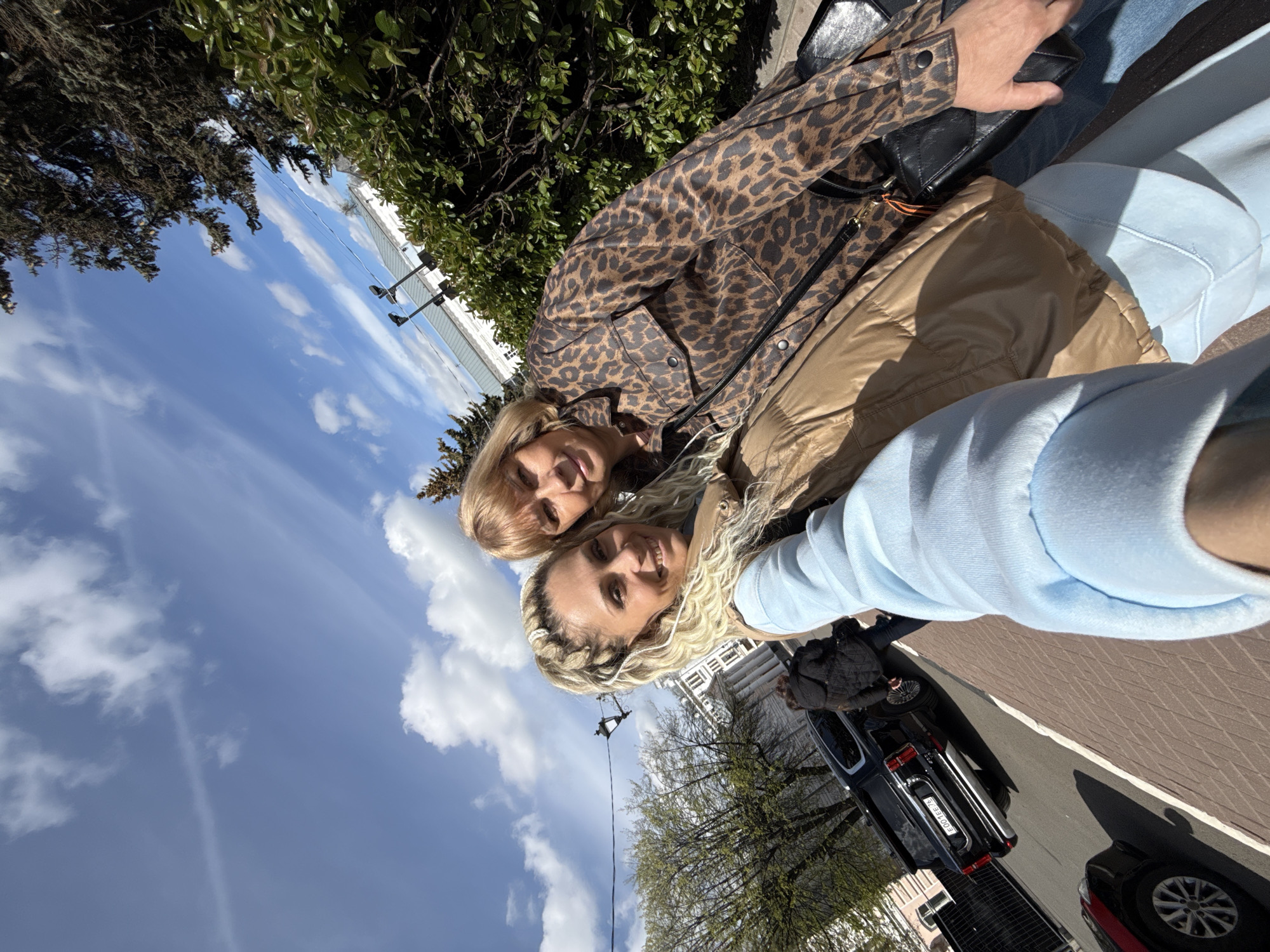 The image shows two women taking a selfie outdoors. The woman in the foreground has blonde hair in braids with a white headband, a light blue hooded sweatshirt, and a light brown puffy vest. The woman behind her has blonde shoulder-length hair, a brown jacket with leopard print, and a black shoulder bag. Behind the women, there are green bushes, trees with new leaves, street lamps, and vehicles parked on a paved street. The sky is blue with white clouds.