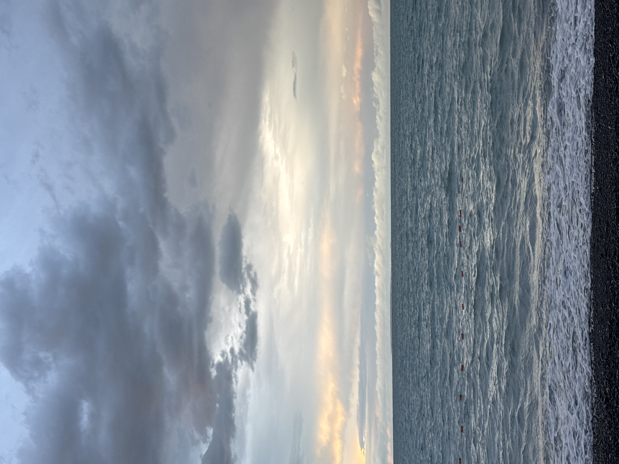 The image captures a view of the water at a beach in the city of Ақши, Kazakhstan. The sky is overcast with dramatic, dark gray clouds. A thin band of soft, pale orange and yellow light is visible near the horizon, suggesting either sunrise or sunset peeking through the clouds. The sea is a deep teal color with visible waves and white foam where they meet the shore. Several small, orange buoys are spaced out in a line on the water's surface. The beach in the foreground is covered in dark, smooth pebbles. There are no people or animals visible in the scene.