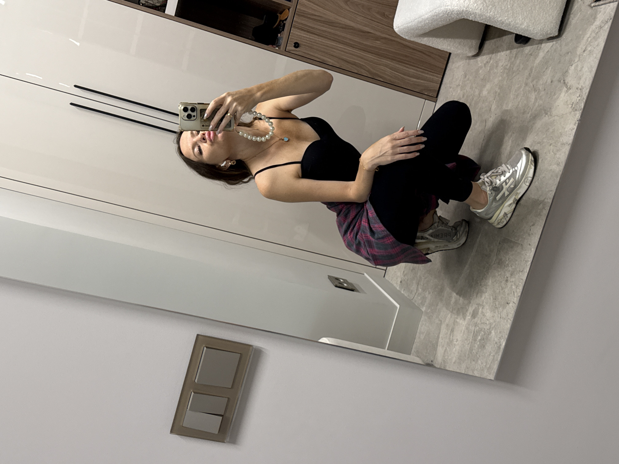 The image shows a young woman taking a mirror selfie in a modern apartment. She is wearing a black tank top, black leggings, and silver sneakers. A pink and black flannel shirt is tied around her waist. She is also wearing a pearl necklace with a blue pendant and has on small hoop earrings. The room appears to be a closet or dressing area with large, white, reflective closet doors. A portion of a wooden cabinet with shelves is visible in the background, as is a fluffy white stool. The floor is tiled with a grey, marbled pattern. The woman is squatting down to get a full-body shot and is holding her phone up to take the picture. Her expression is playful, with her lips pursed. The overall lighting suggests it is daytime, likely indoors. The presence of the closet doors and a stool suggests a private dressing or grooming space. The location context provided is Khimki, Russia. There is no visible text in the image.