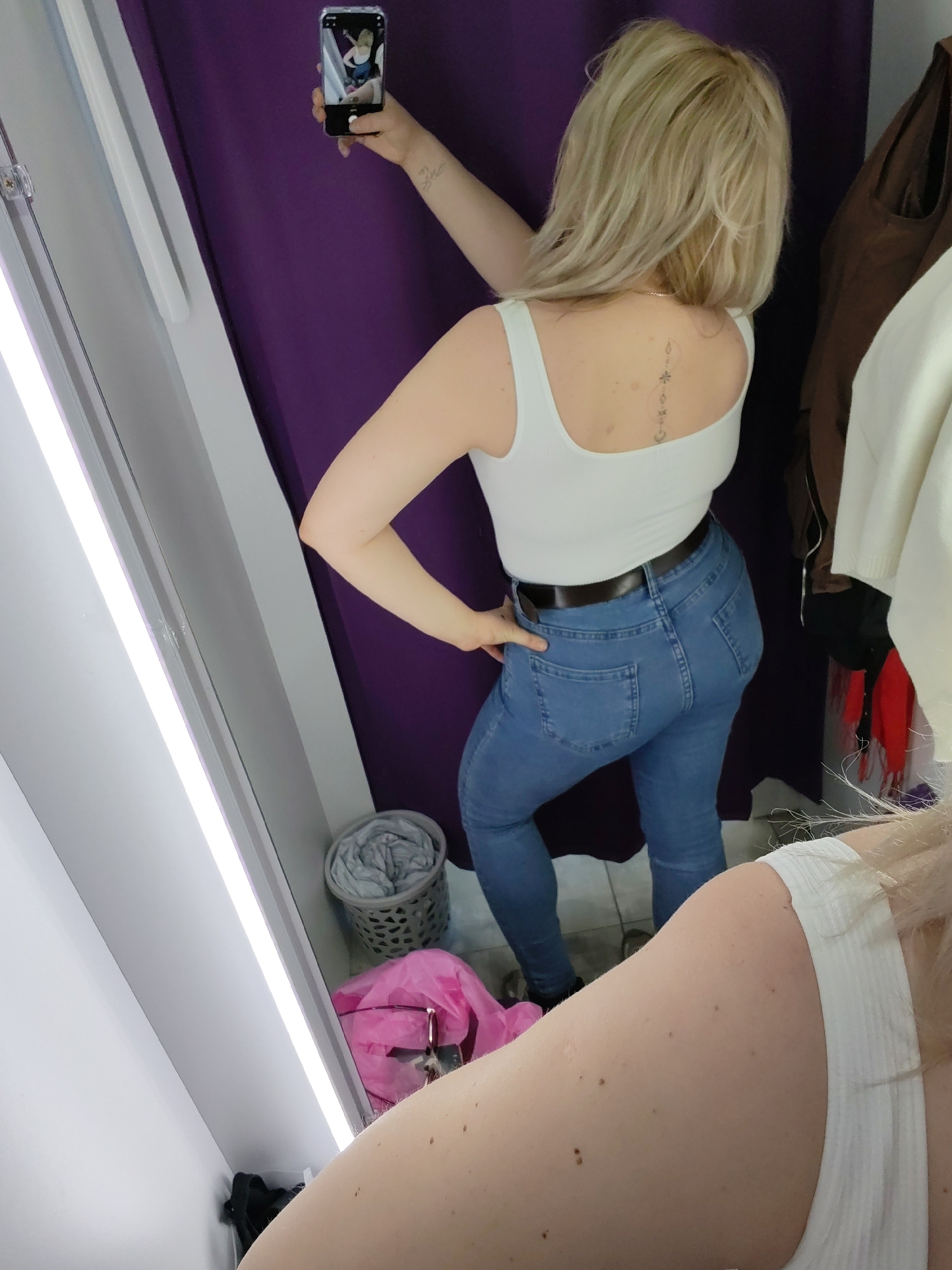 A blonde woman in a white tank top and blue jeans with a brown belt is taking a selfie in a dressing room. The dressing room has a purple curtain, a white wall, and a mirror with a light on the side. There's a grey basket and a pink garment on the floor. The woman is holding a smartphone to take a photo of her reflection in the mirror. The woman has a tattoo on her back. The location is not discernible as Izhevsk, Russia.