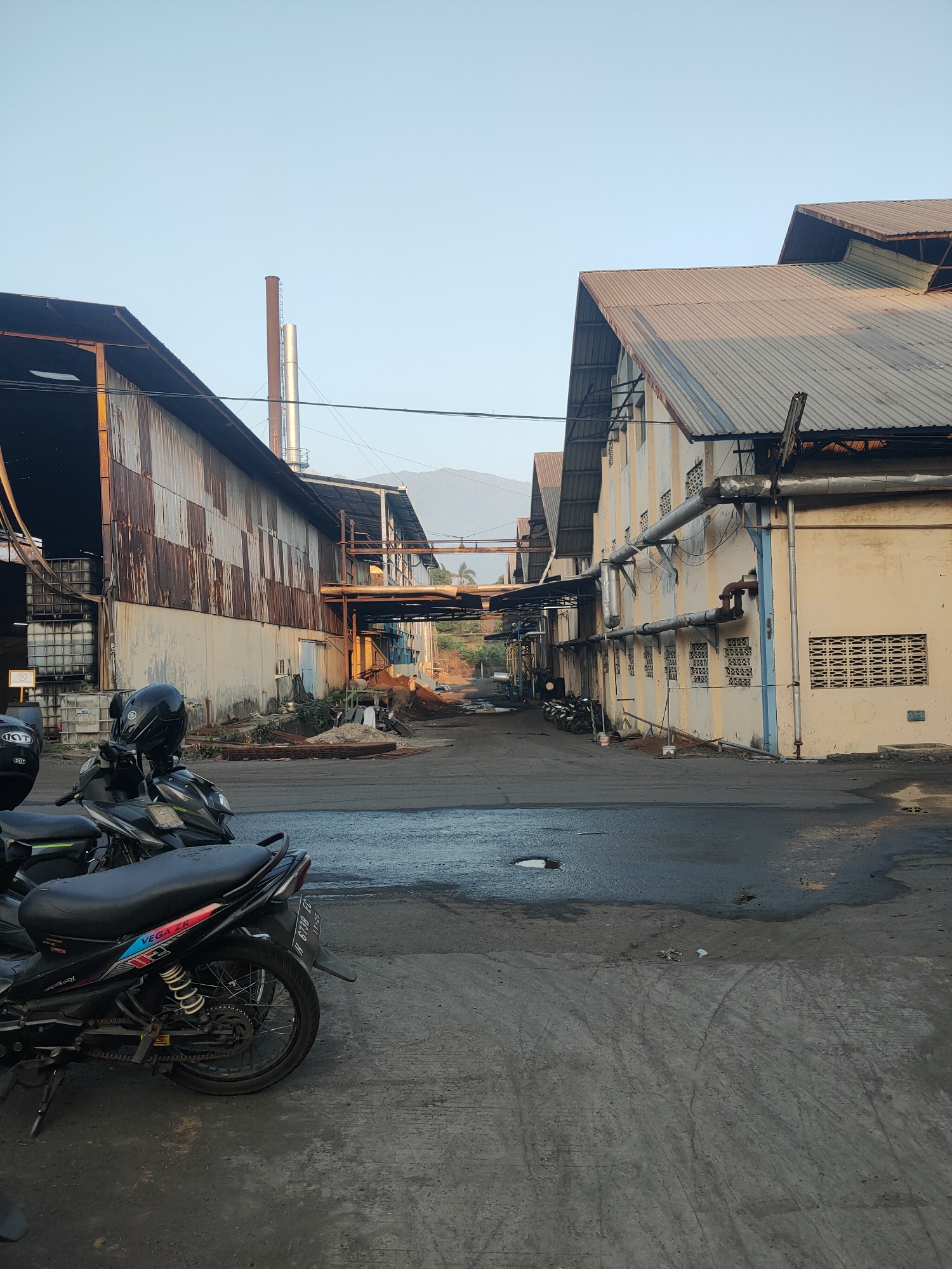 The image depicts an industrial area with two large buildings, one on the left with a corrugated metal facade showing signs of rust, and another on the right with a yellow painted exterior and a corrugated metal roof. Both buildings have industrial pipes running along their exteriors. A tall chimney stack is visible behind the building on the left. The ground is paved with asphalt, and there are puddles of water and debris scattered around. Several motorcycles are parked in the foreground and along the side of the building on the right. The sky is clear and pale blue, with a distant view of hills or mountains. The scene appears to be in an industrial zone, possibly a factory or warehouse complex, located in the city of Beji, Indonesia.
