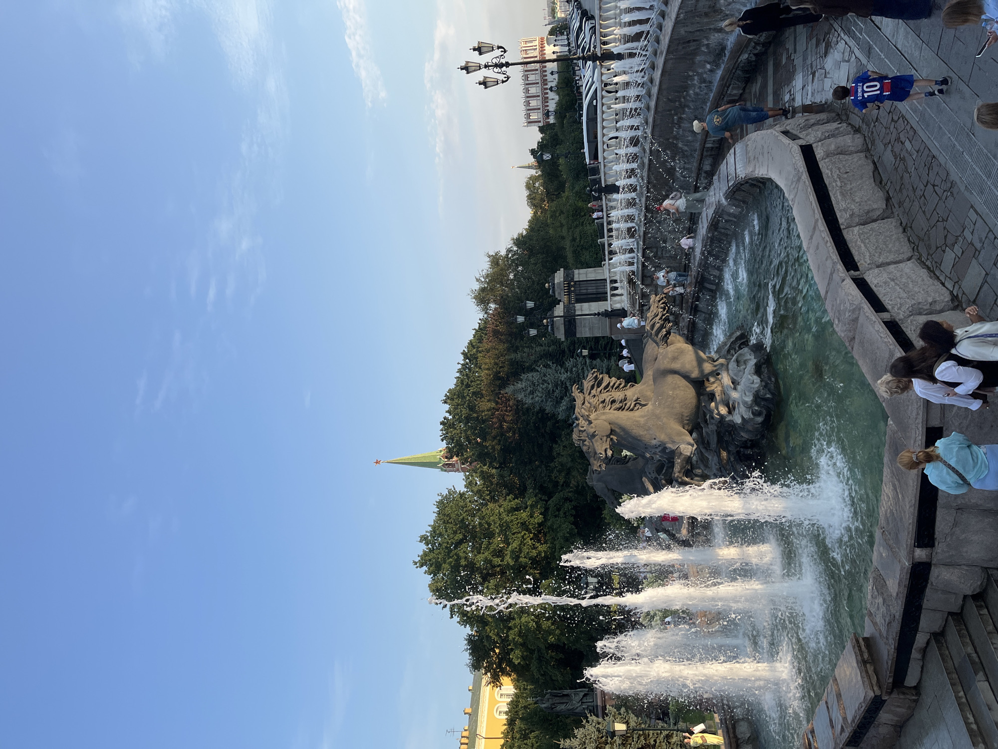 The image shows a view of the Alexander Garden in Moscow, Russia, with a large fountain in the foreground. The fountain features a sculpture of horses. Several people are visible around the fountain and on the pathway above it, some of whom are walking and others observing. In the background, there are trees, a building with red and white accents, and a green spire with a red star, likely a Kremlin tower. The sky is blue with scattered clouds. The lighting suggests it is daytime, possibly late afternoon or early evening.