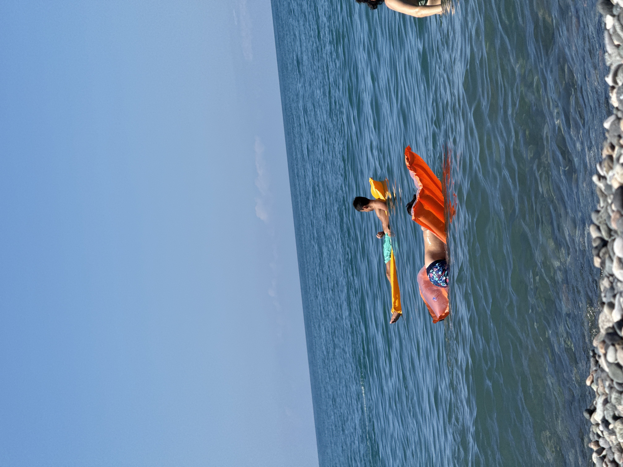 An eye-level, medium shot captures a serene beach scene under a clear blue sky. In the foreground, a textured expanse of grey and white pebbles lines the water's edge, hinting at a rocky shoreline. The deep blue sea, with gentle ripples on its surface, dominates the middle ground and background. Three individuals are visible, each enjoying the water on inflatable mattresses.
Closest to the shore, an individual is lying on an orange inflatable mattress, their lower body visible, clad in patterned swim trunks. Further out, a second person rests on a yellow inflatable mattress, their body facing away from the camera, wearing teal swim shorts. In the distance, a third individual, mostly out of frame on the right, appears to be standing in the water. The overall impression is one of peaceful leisure and a summer day spent by the sea.