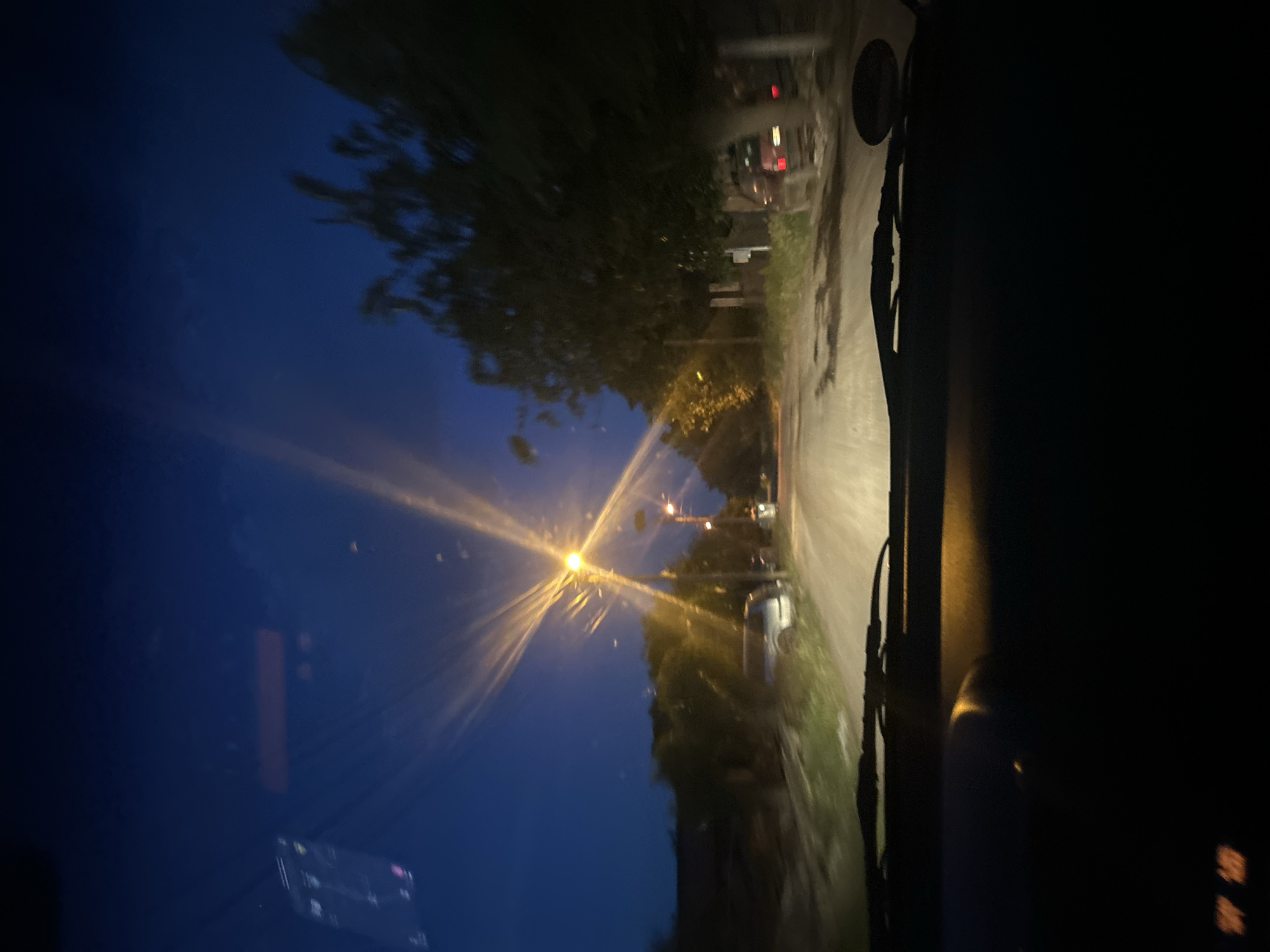 The image shows a view from inside a vehicle, looking out onto a road at night. A street lamp casts a bright glow onto the road and into the sky. Buildings and trees line the road. The road appears wet.
There is not enough information in this image to determine the location in the city of Pishtigovo, Bulgaria.