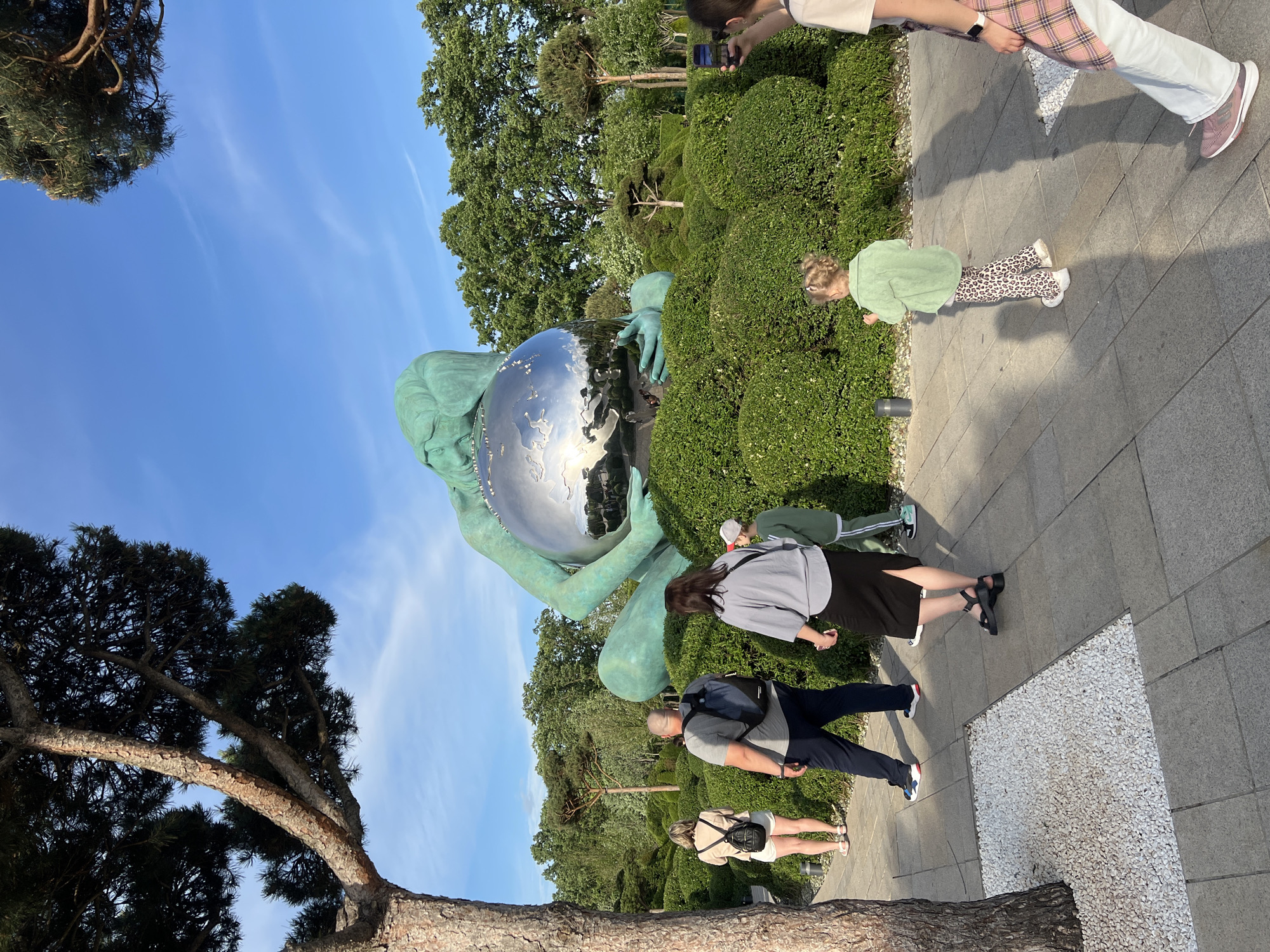 A bronze statue of a woman holding a silver orb is in a park setting in Krasnodar, Russia. The statue sits among green shrubbery. Several people are in the vicinity of the statue on a gray paved walkway. A child in a green jacket and leopard-print pants is near the shrubbery. A woman in a gray sweatshirt and black skirt stands on the walkway. An older man in a gray shirt and navy pants is also on the walkway. A woman wearing a tan top and white shorts is walking away from the statue. A person near the top of the frame is taking a picture with a smartphone. A tree is in the lower left corner of the frame. The sky is blue with some clouds.