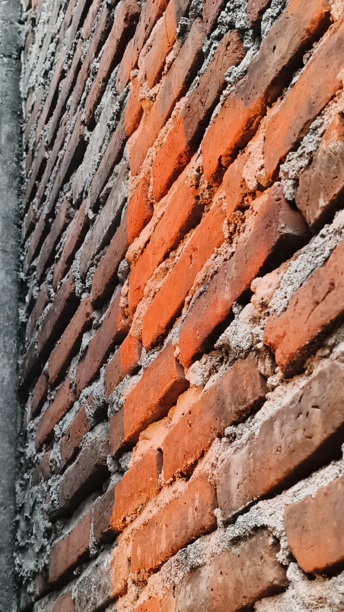 The image shows a close-up, angled view of an unfinished brick wall. The bricks are predominantly reddish-brown with rough textures, and they are laid with visible gray mortar. A portion of the wall is illuminated by a warm orange light, highlighting the texture and color of the bricks. To the left, a grey concrete structure, possibly a pillar or edge of another building, runs vertically along the side of the brick wall, contrasting with the brickwork. The scene appears to be an outdoor setting, possibly under construction or in a rustic environment. The location is identified as Kerep Kidul, Indonesia. There are no people or animals visible in the image, and no specific actions or interactions are depicted. The focus is entirely on the architectural elements of the wall.