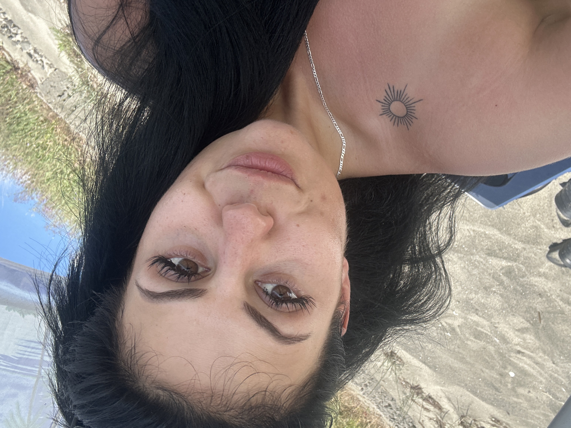The image is a portrait of a woman with long, dark hair, taken outdoors. Her face is the primary subject, with her eyes open and looking towards the camera. She has dark eyebrows and long eyelashes. Her lips are slightly parted, and she appears to be wearing subtle makeup. Around her neck, a thin silver chain is visible.
On her upper chest, slightly to the right of her collarbone, there is a small tattoo of a sun with radiating lines. The skin in this area shows some minor texture or stretch marks. Her hair is dark and appears to be in waves, some strands falling across her face and shoulder.
The background suggests an outdoor setting, with elements of ground and possibly a walkway or paved area visible. There are also hints of green vegetation and a bright blue sky, indicating it is daytime with likely clear or partly cloudy weather. The overall impression is a casual, personal photograph, possibly a selfie, taken in a relaxed environment. The lighting suggests natural sunlight. No visible text is present in the image. The orientation of the image is rotated.