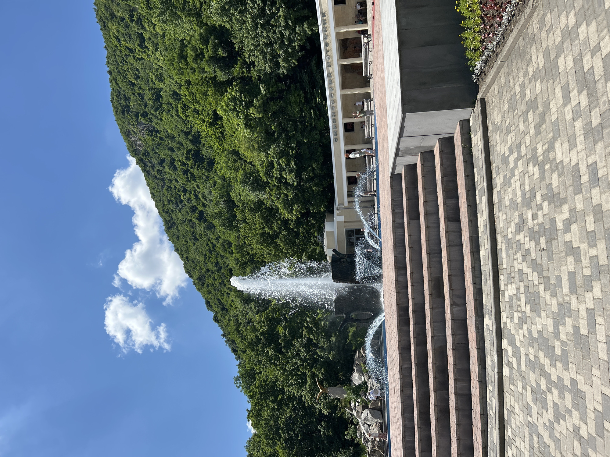 In Zheleznovodsk, Russia, this image captures a wide shot of a public space featuring a large fountain as its central element. The fountain, constructed from dark, textured material resembling large vessels, sprays water high into the air, with smaller streams and splashes reaching outward. To the left of the fountain, a statue of a golden eagle is perched on a rocky outcrop, with a small group of people observing the scene. Behind the fountain and people, a white, colonnaded building is visible, with what appears to be Cyrillic text above its entrance. The building is set against a backdrop of a lush, green mountainside densely covered in trees. In the foreground, a wide, multi-tiered set of stairs made of brick and stone leads up to a paved area. The sky above is a clear blue with a few scattered white clouds. The paving stones in the foreground are in a two-tone patterned arrangement. The overall scene suggests a park or recreational area, possibly a tourist attraction, in a mountainous region.