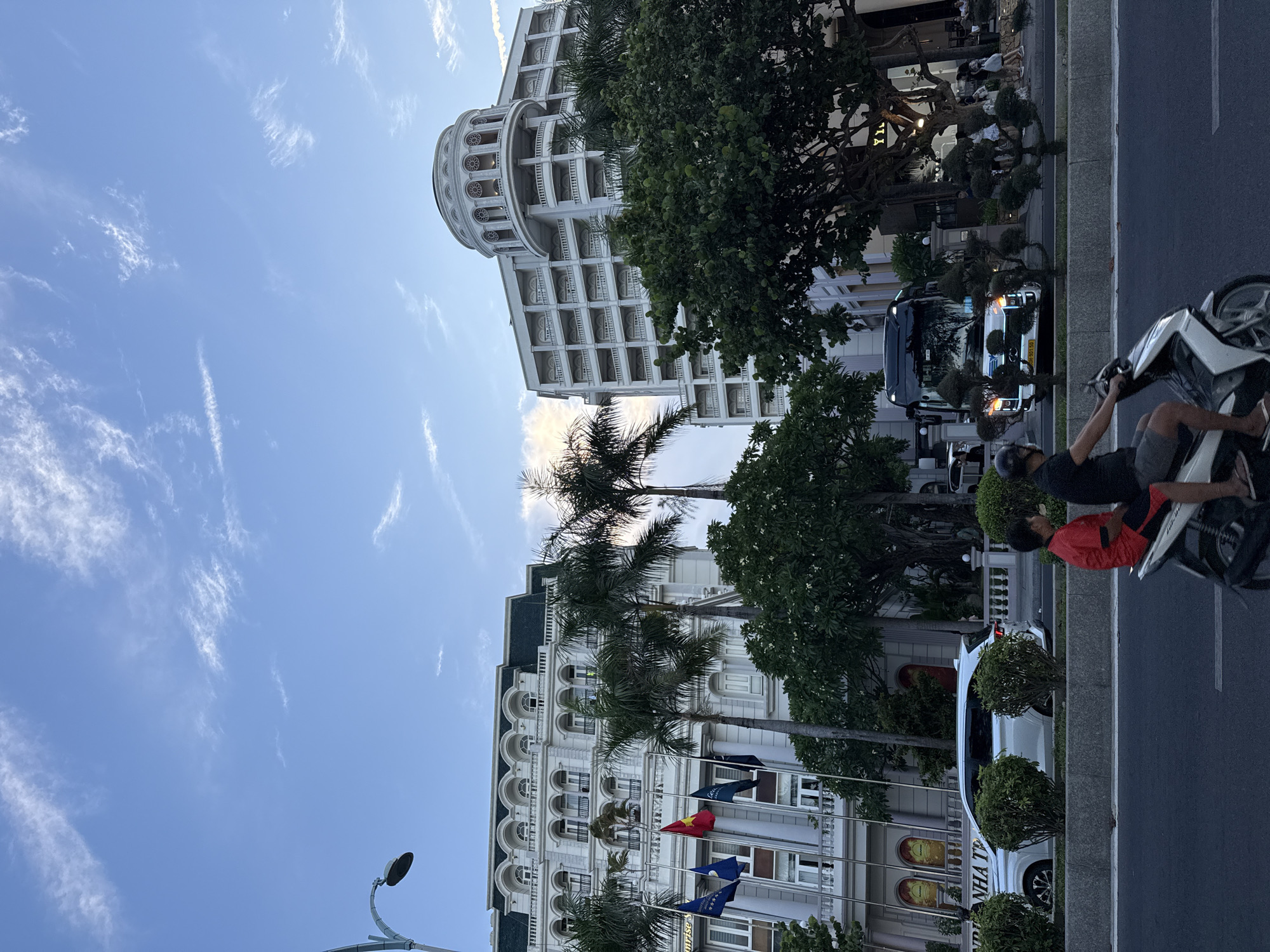 In Nha Trang Ward, Vietnam, a street scene unfolds under a partially cloudy sky. Two large, white buildings with ornate architecture dominate the background, one featuring a prominent dome. In the foreground, a paved road is occupied by a scooter with two male riders, one wearing a red shirt and black shorts, the other a black shirt and gray shorts. They are positioned to the left of the frame, moving away from the viewer. To their right, a white SUV is parked, and further down the street, a black bus is visible. Palm trees and lush green foliage line the sidewalk, interspersed with decorative, sculpted bushes. A lamppost stands on the left edge of the image. People are visible walking on the sidewalk in the distance. The overall impression is one of urban activity in a tropical setting.