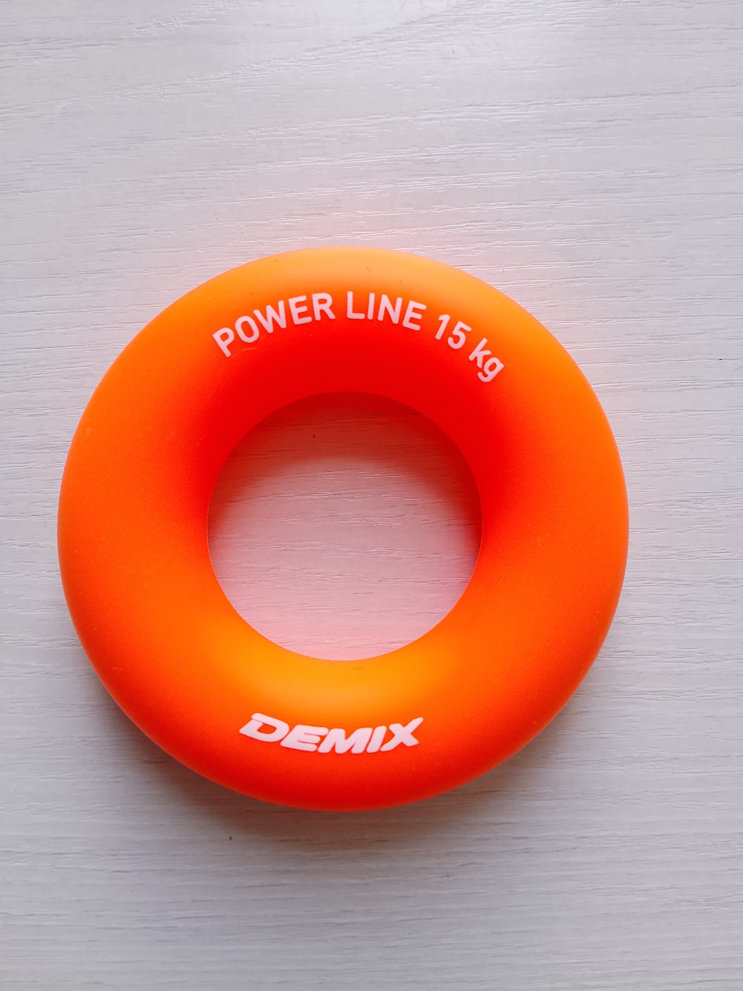 An orange-colored, ring-shaped hand exerciser lies on a white, textured surface. The hand exerciser has white lettering on it. On the top curve of the ring, the text "POWER LINE 15 kg" is visible. On the bottom curve of the ring, the word "DEMIX" is visible. The background is a light-colored, horizontal striped texture, possibly a wooden surface. The image is taken from a top-down perspective, centered on the hand exerciser. There are no people present, and no actions or interactions are depicted. The location is not specified within the image.