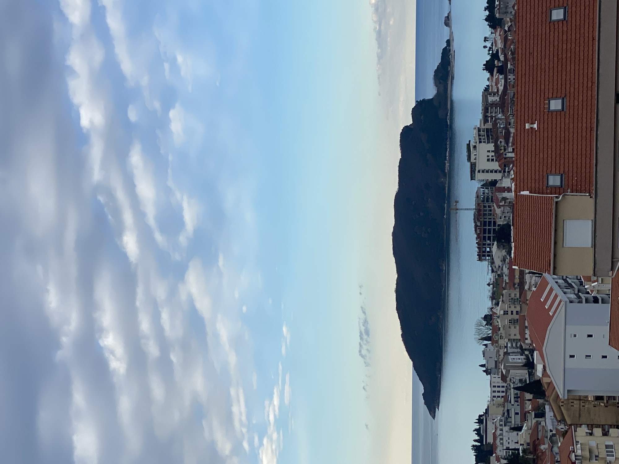 The image depicts a cityscape and seascape, likely taken from an elevated vantage point. The foreground consists of building rooftops with red tile roofs and white facades, typical of Mediterranean architecture. The midground features a body of water, possibly the Adriatic Sea, and a dark, mountainous landmass extending into the water. In the distance, more buildings are visible along the coastline. A construction crane is visible, indicating ongoing development. The sky is partly cloudy with a mix of light blue and gray tones. The overall scene suggests a coastal urban area in Budva, Montenegro, characterized by its mountainous coastline and clustered architecture.