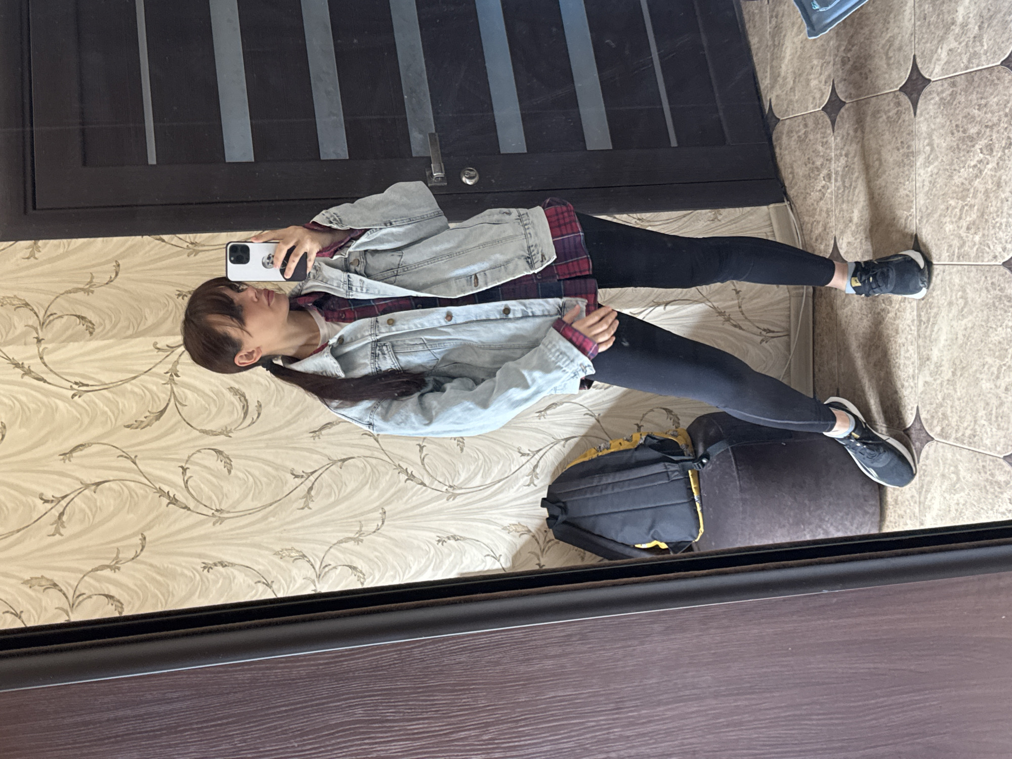 A full-length reflection shows a person in dark leggings, a red plaid shirt, and a light denim jacket. They are holding a white smartphone. A black backpack with yellow accents is on a dark-colored ottoman. The reflection is captured in a mirror with a dark brown frame. Behind the person, the wallpaper is a light beige with a flowing, floral pattern. The flooring is tiled in varying shades of light brown. The door behind the person is dark with silver inlays. The location is an indoor setting, likely in Rostov-on-Don, Russia.