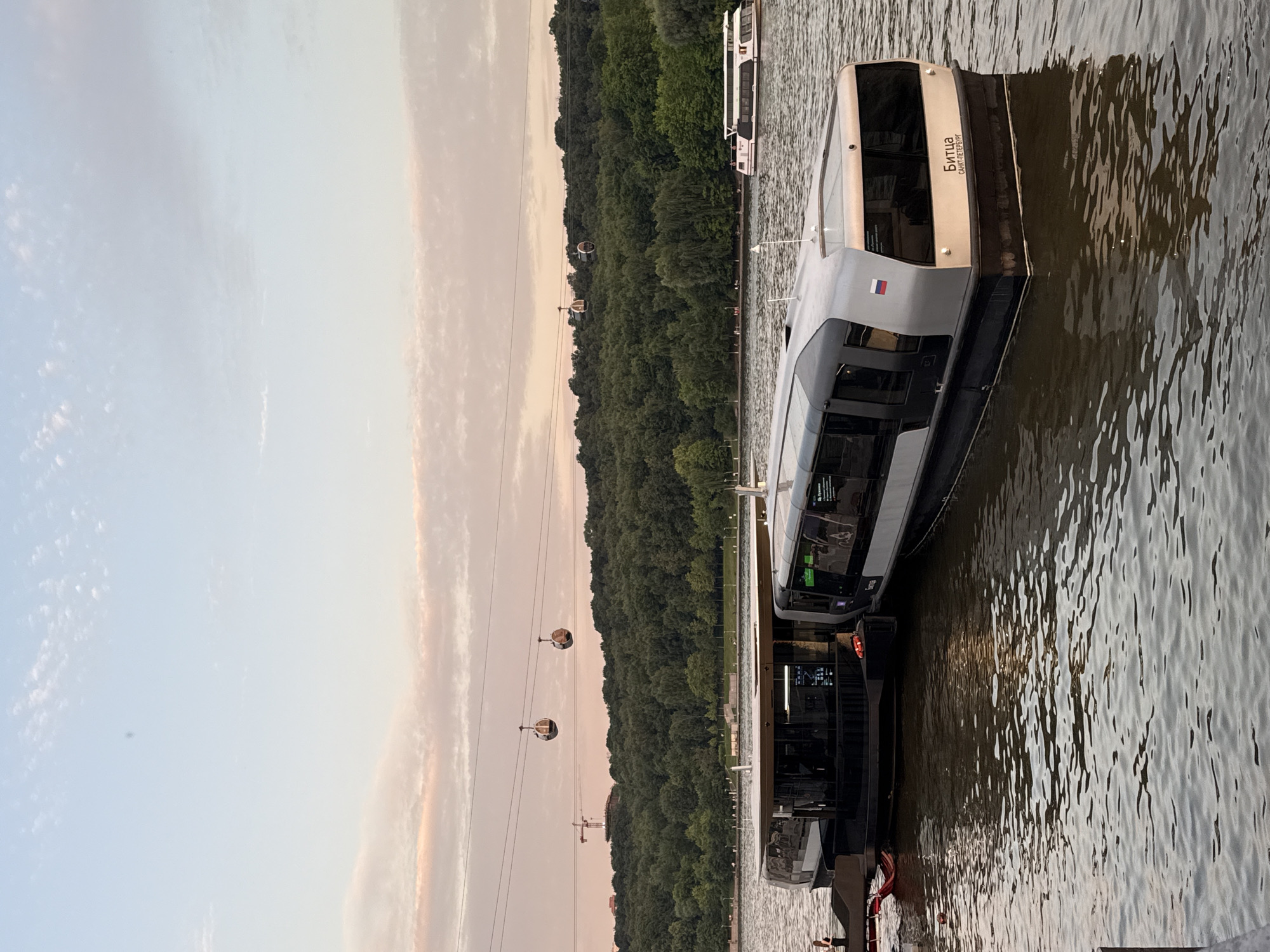 This image captures a view of the Moscow River in Moscow, Russia, likely during the evening or sunset given the soft, pastel hues in the sky. In the foreground, two modern, sleek passenger boats are docked side-by-side. These boats are predominantly gray and black, with large, tinted windows. The boat on the right has Russian text on its stern, reading "Битца" and "САНКТ-ПЕТЕРБУРГ."
In the middle ground, the river stretches out, reflecting the sky and the surrounding greenery. Across the river, a lush, dense forest covers a hill. Above the treeline, two cable car cabins are suspended on wires, suggesting a cable car system operating in the vicinity. A construction crane is visible in the far distance, behind the trees and to the left of the cable cars, indicating ongoing development or infrastructure projects in the city. Another, larger white passenger boat is visible further down the river to the right. The water exhibits a gentle ripple, and the overall atmosphere is serene and picturesque.