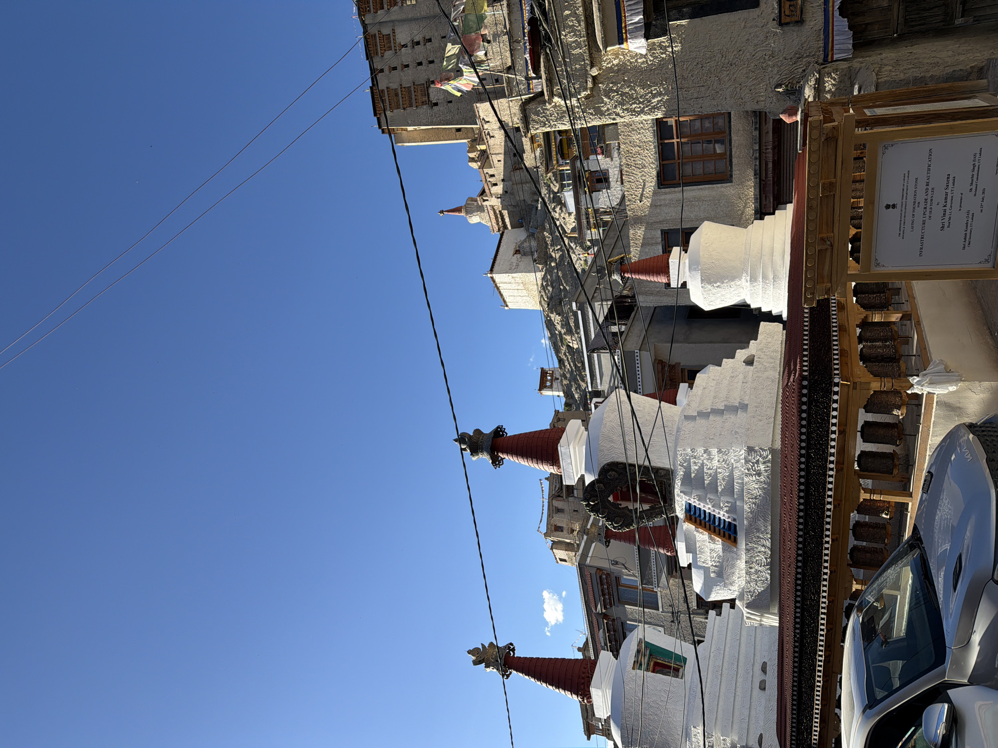 The image depicts a scene in Leh, India, likely at a Buddhist monastery or cultural site. In the foreground, a silver car is partially visible. Behind the car, a wooden structure with prayer wheels is prominent. Several white stupas, characteristic of Buddhist religious architecture, are arranged in the mid-ground. These stupas have red conical tops adorned with golden finials. Further in the background, a large, multi-story fortress-like structure is built on a hillside. The architecture of the buildings suggests a traditional Ladakhi style, with earthen and stone construction. Prayer flags are visible, fluttering in the breeze on some of the structures. The sky is clear and blue, indicating a sunny day. A sign board is also visible in the lower right corner, providing information about an infrastructure upgrade and beautification project. The overall scene conveys a sense of cultural heritage and spirituality in a mountainous region.