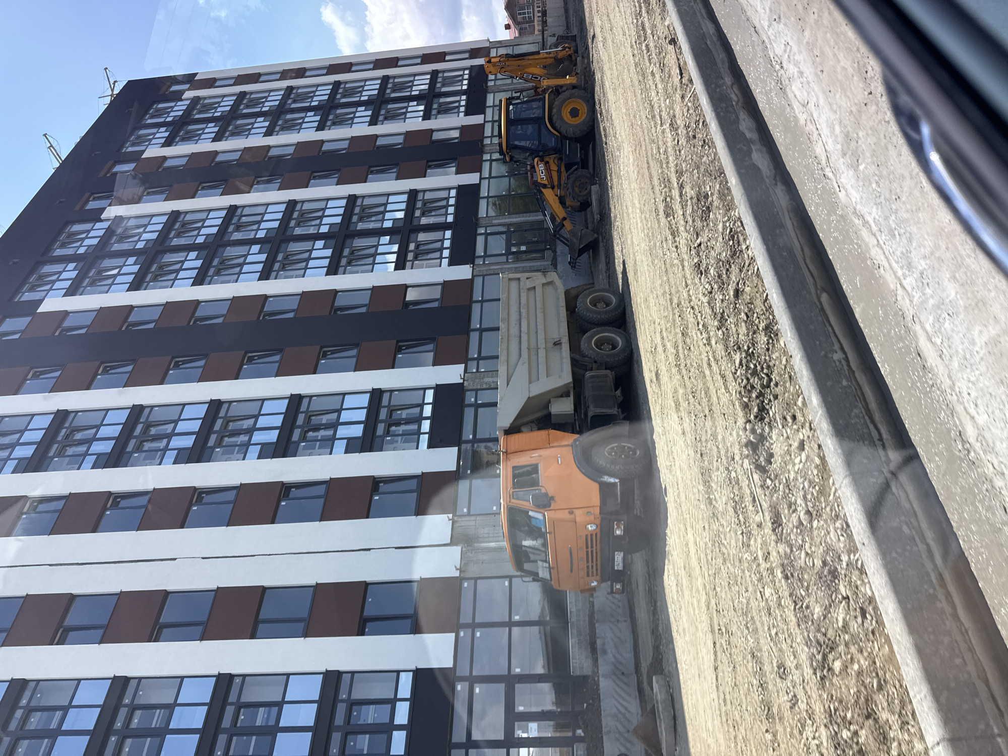 In Mikhaylovsk, Russia, on a bright day, a construction site is visible featuring a tall, modern apartment building. The building has a distinctive facade with alternating vertical bands of white and brown, interspersed with dark windows. In front of the building, heavy machinery is present. An orange dump truck is positioned on the left side of the frame, and a yellow backhoe loader, identified by the "JCB" logo, is on the right. Both vehicles are on a dirt and gravel surface, suggesting ongoing construction work. The ground is uneven and covered with debris typical of a work zone. A concrete curb is being formed in the foreground, with wet concrete visible. The sky above is partly cloudy. No people are discernible in the image.