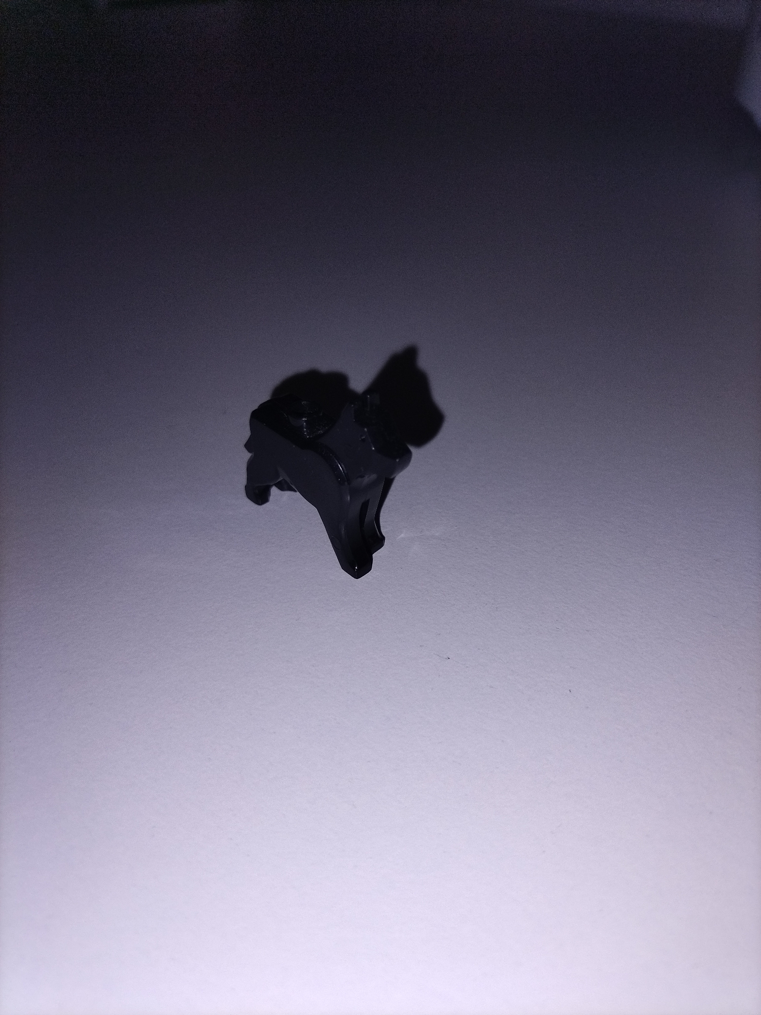 A small, dark-colored figurine, resembling an animal like a horse or a dog with a saddle or rider element on its back, is the central subject of the image. The object appears to be a toy or game piece, possibly made of plastic. It is positioned on a flat, light grey or off-white surface that displays a faint, uniform texture. The background is dark, transitioning from deep grey or black at the top to a lighter grey closer to the foreground.
The scene is dimly lit, with the primary light source appearing to come from the front and slightly to the right of the figurine. This lighting creates a distinct shadow that extends to the left and behind the object, enhancing its silhouette. The dark object contrasts sharply with the lighter surface it rests upon. There are no people present, no specific activity is taking place beyond the static display of the object, and no text is visible. The low light conditions contribute to a somewhat muted and focused atmosphere. No specific location, time of day, or weather conditions can be discerned.
