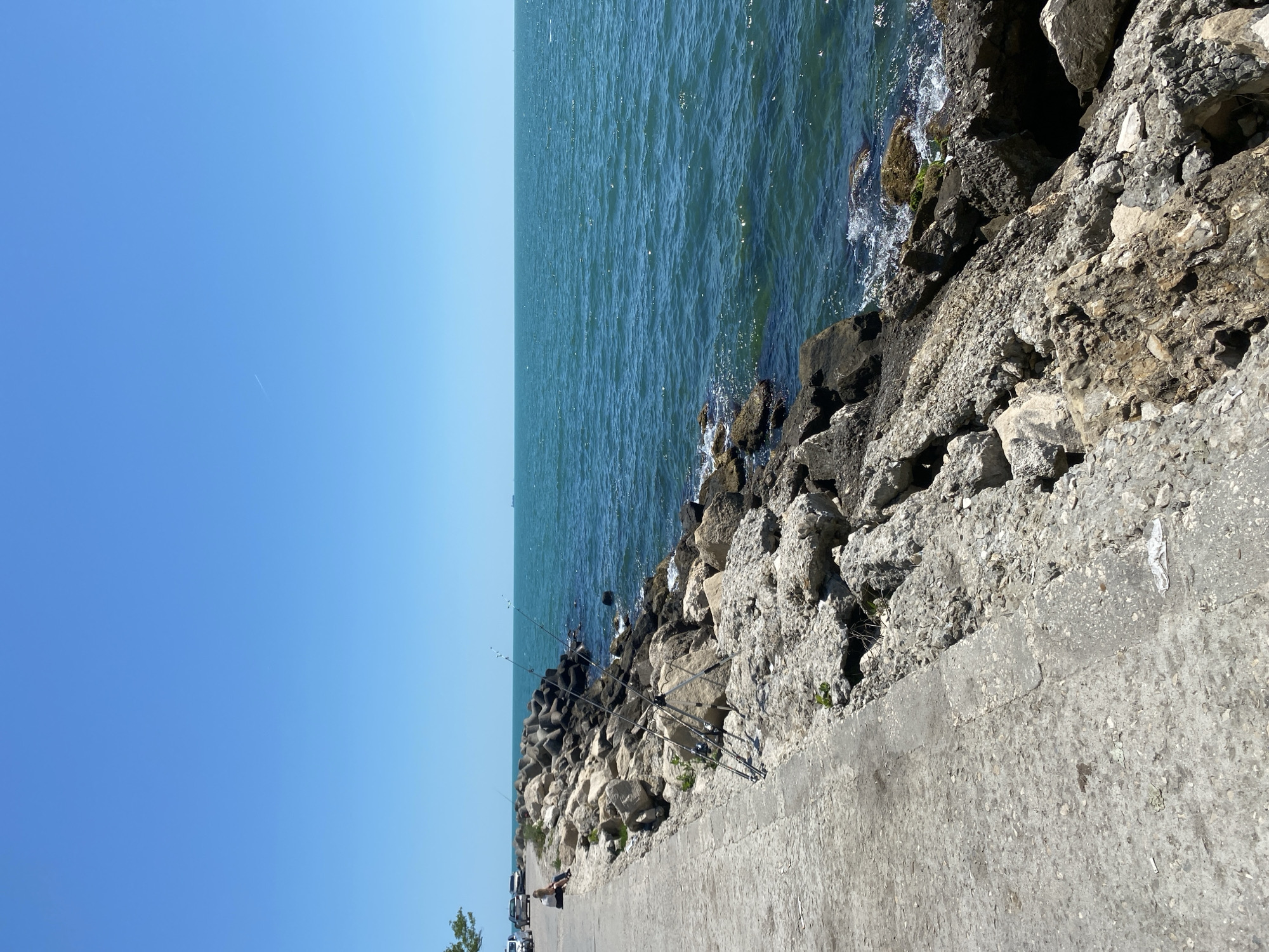 The image depicts a seaside scene in Varna, Bulgaria, likely on the South Beach promenade or a nearby pier. The foreground features a concrete walkway bordering a rocky shoreline. Several fishing rods are set up on the rocks, suggesting active fishing. A person is seated on the walkway, facing the sea, with cars parked on the left side of the frame, indicating a public area. The sea is a vibrant blue-green, with gentle waves lapping against the rocks. The sky is clear and a bright blue, with a faint contrail visible in the upper right quadrant. The overall impression is of a tranquil day at the coast.