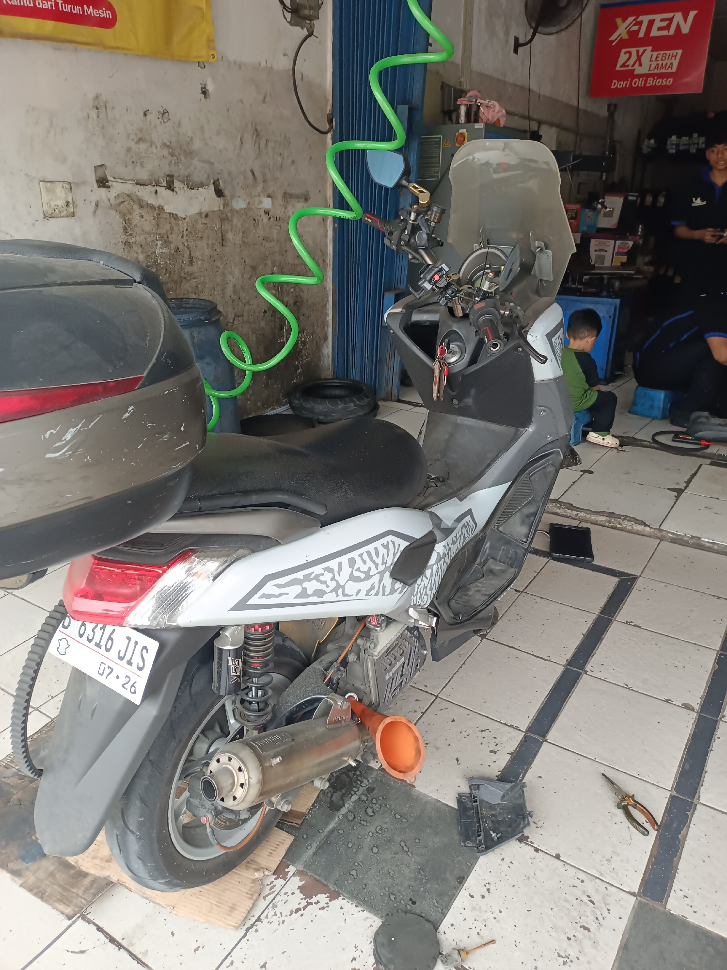 This is a medium shot of a motorcycle repair shop in Serdang Wetan, Indonesia. In the foreground, a white and silver scooter is parked on a cardboard mat. The scooter has a black seat, a windshield, and a silver exhaust pipe. An orange funnel is placed on the ground near the scooter, along with a pair of pliers and some other tools. In the background, two young men are visible. One, wearing a green shirt, sits on a blue stool. The other, in a blue shirt, stands behind a counter, holding a tablet. The shop has a tiled floor and a blue metal shutter door. A green hose hangs from the ceiling. A yellow banner with Indonesian text is displayed above the workspace.
