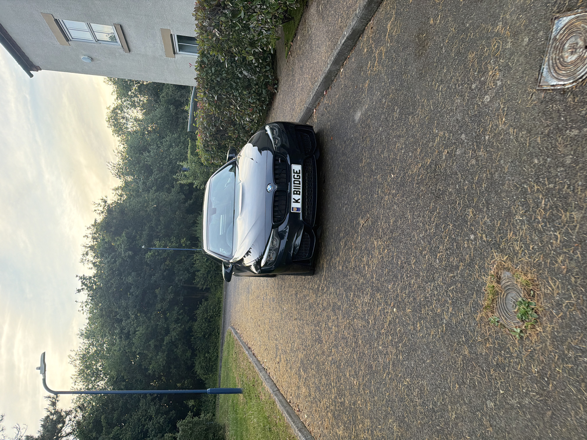 A black BMW M4 Coupe, bearing the UK registration plate 'K BIIDGE', is prominently positioned on an asphalt road in Maidstone, United Kingdom. The vehicle is angled slightly towards the viewer, facing directly forward. The asphalt surface is largely obscured by a dense layer of light brown, needle-like plant debris.
To the left of the car, a concrete curb delineates the road from a strip of green grass. Beyond the grass, a tall blue street lamp with a curved neck and grey fixture stands erect. A dense line of green trees and undergrowth forms the background on this side, with a second, thinner blue pole barely visible further down the path.
To the right, a parallel concrete curb separates the asphalt from a narrow, lighter-toned paved path. Behind this path, a well-maintained hedge with dark green and some reddish leaves partially conceals a light-grey rendered two-story building. The visible portion of the building features two white-framed windows, one with a prominent brown sill, and a circular wall-mounted fixture between them.
The sky overhead is overcast, displaying soft, warm, yellowish-white light, indicative of either late afternoon or early evening. Two circular metal utility covers are partially visible embedded in the asphalt, with one in the immediate foreground showing minor weed growth. No individuals or animals are present in the scene.