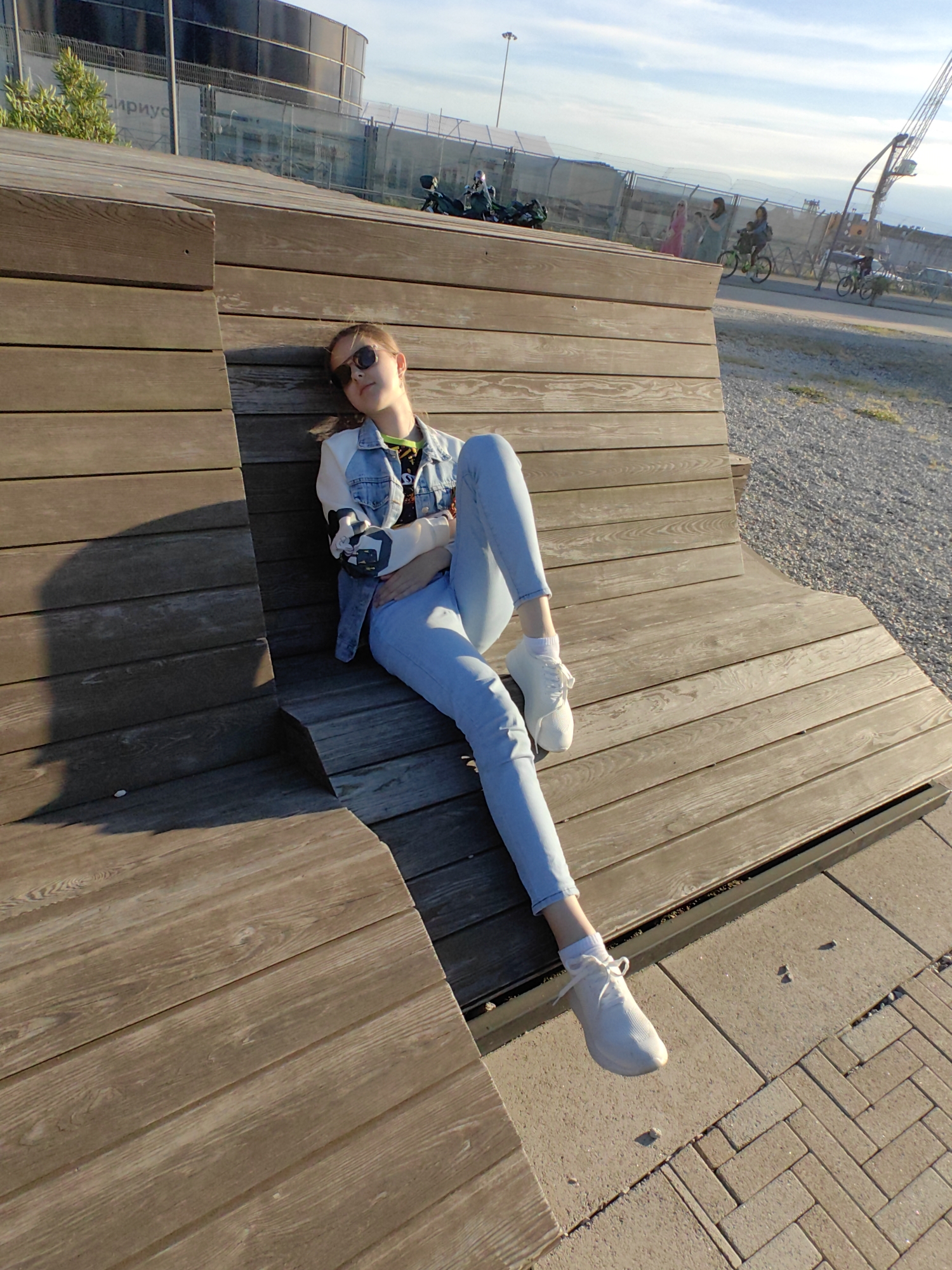 This eye-level outdoor photograph captures a young female relaxing on a large, multi-level wooden bench bathed in warm, late afternoon sunlight, indicative of the golden hour. The sky above is clear with scattered clouds.
The main subject, positioned on the left side of the frame, is casually seated, leaning back against the horizontally slatted wooden structure. She wears dark sunglasses, a light blue denim jacket with graphic designs on the sleeves, a bright green or yellow top, light blue jeans, and white sneakers with white socks. Her posture conveys a sense of comfort and relaxation.
The wooden seating structure, robust and modern in design, dominates the foreground and midground. The ground to its right is a mix of pebbles and light-colored paving stones laid in a herringbone pattern. In the background, a chain-link fence delineates the leisure area from a public thoroughfare. Behind this fence, several other people are visible engaged in various activities: two individuals on motorcycles or scooters, three pedestrians including one in a vibrant pink dress, and two people cycling further down the path.
Further in the distance, a large, dark, cylindrical building is prominent, partially obscured by fencing and some sparse greenery. The Cyrillic text "ИРИУС" is partially visible on this structure, confirming the location as Sirius, Russia. A tall, thin metal pole or structure also extends upwards towards the sky. The scene depicts a lively yet relaxed urban outdoor environment, likely a promenade or waterfront area, where people are enjoying a sunny day.