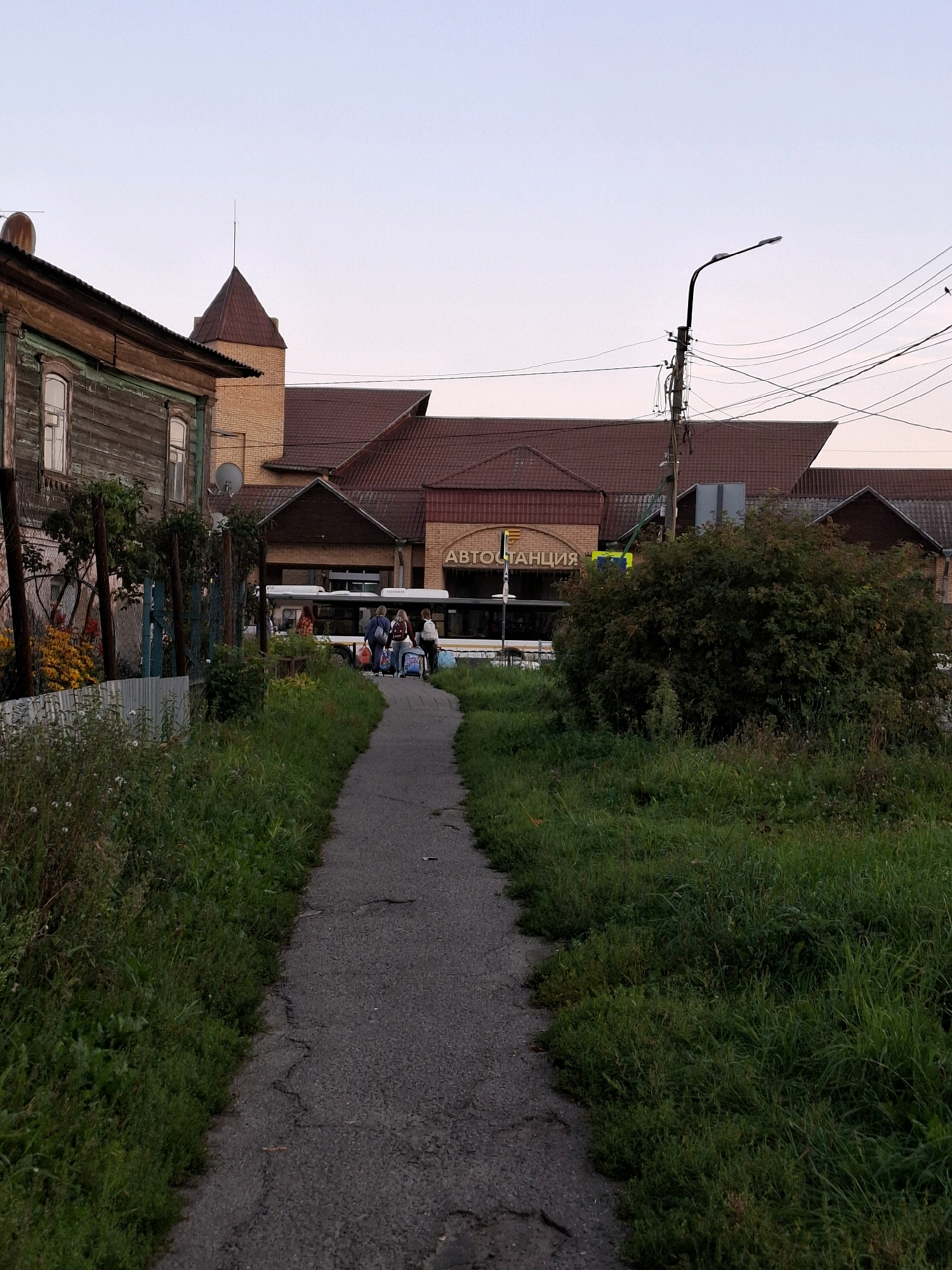 The image depicts an outdoor scene, possibly a bus station, in an urban environment. In the foreground, a cracked asphalt pathway leads towards a building with a red-tiled roof. Dense green grass lines the path on both sides, with some wildflowers visible on the left.
To the left of the pathway, a weathered wooden building with green trim and windows stands. Behind it, a modern brick building with a prominent red roof and a turret-like structure is visible. A bus is parked in front of this building, and several people, likely passengers, are standing with luggage. The word "АВТОСТАНЦИЯ" (Autostantsiya, meaning Bus Station) is visible on the facade of the brick building.
A street lamp with multiple wires attached to a pole stands on the right side of the image. A large, dense green bush is situated in the mid-ground, partially obscuring the bus station building. The sky is a pale, overcast hue.
