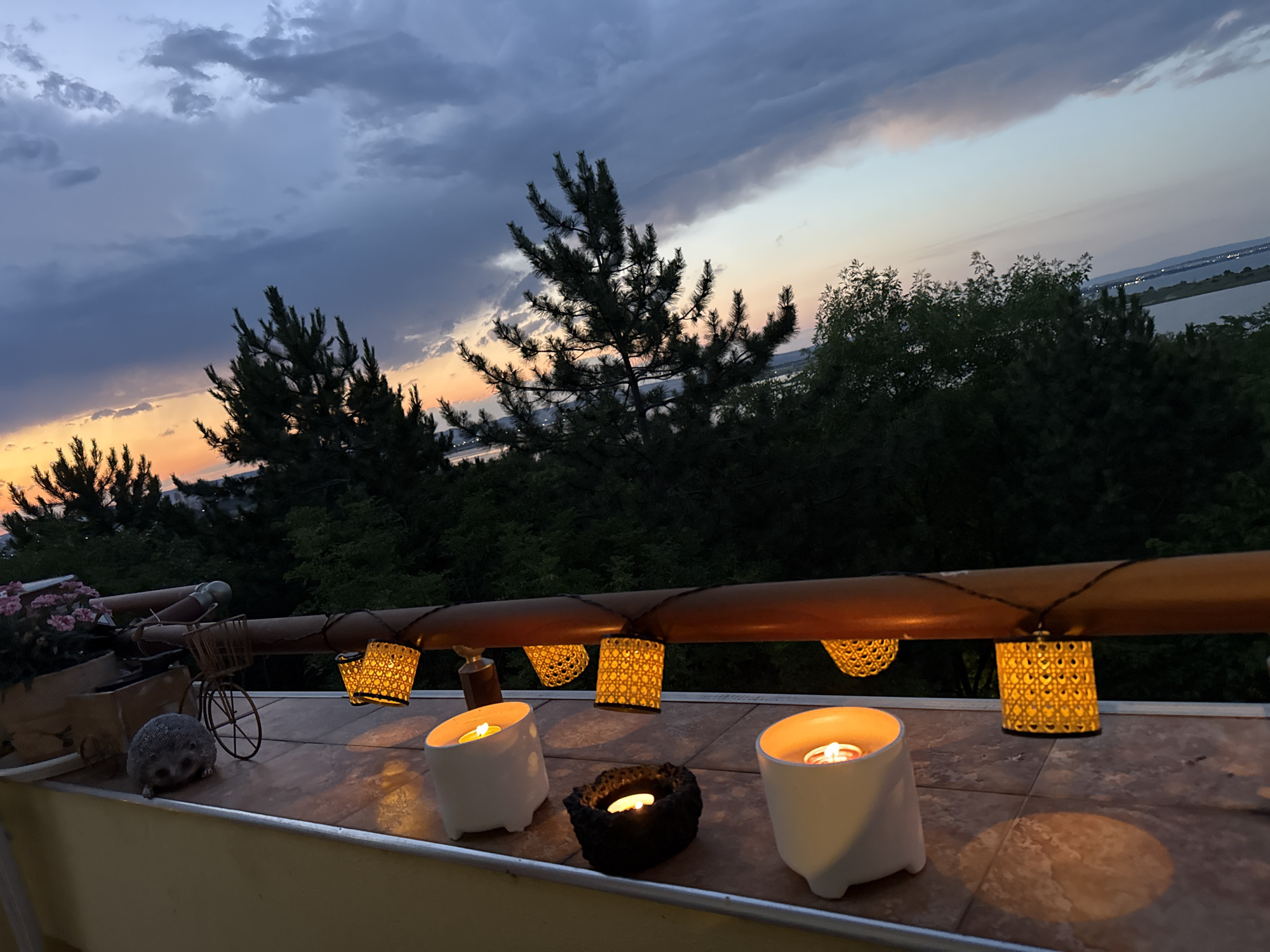 This is a photograph of a balcony overlooking the sea at dusk in Burgas, Bulgaria. In the foreground, there are several lit candles in white and black holders on a tiled surface. A string of decorative lanterns hangs from a wooden railing above. In the background, trees and a body of water are visible, with the sky displaying shades of blue, gray, orange, and pink from the setting sun.
