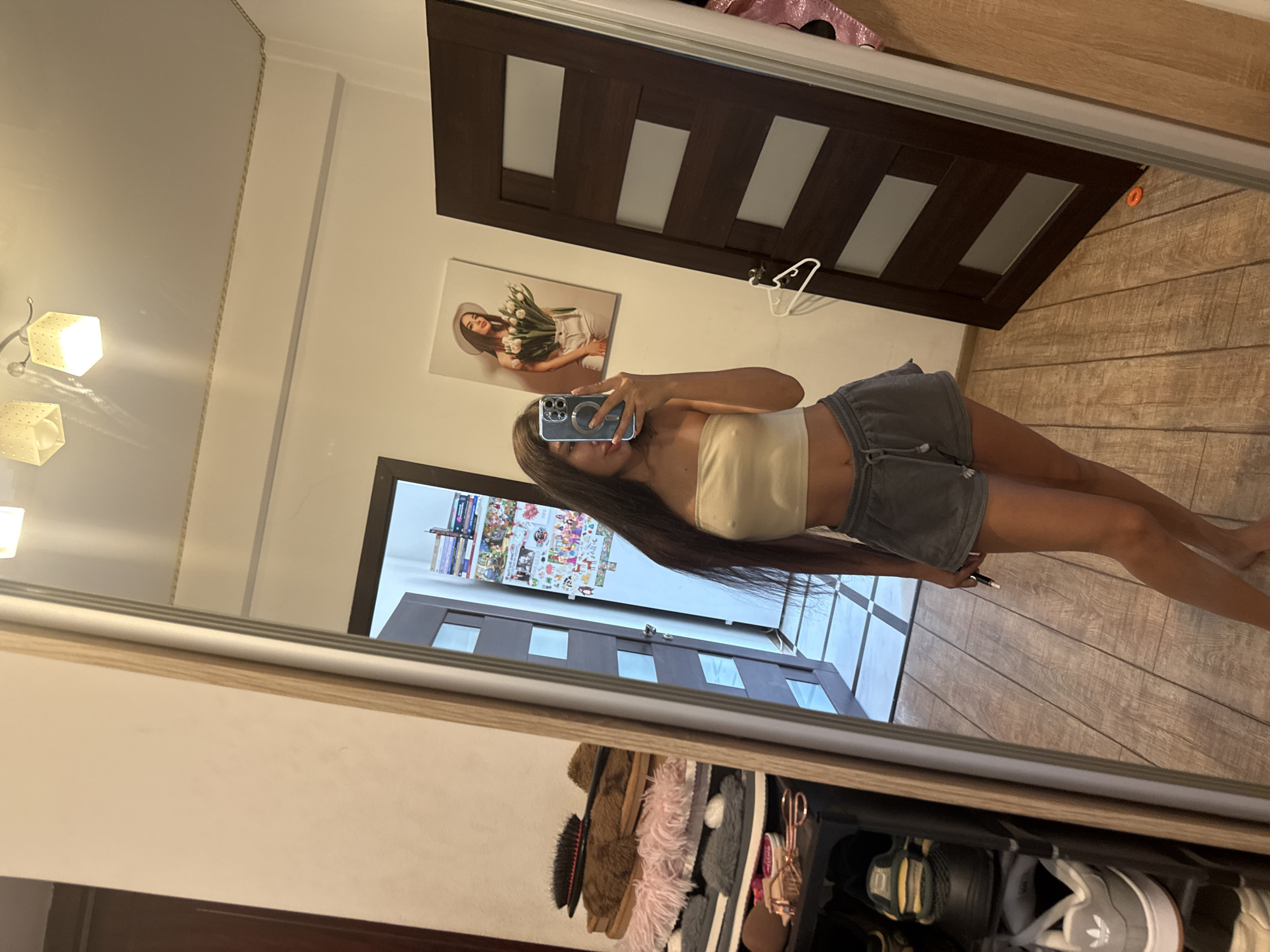 The image shows a young woman in her bedroom, taking a mirror selfie. She is wearing a white bandeau top and gray shorts. In the background, there is a closet with shoes and clothes, a door, and a painting on the wall. The floor is made of wood. The lighting in the room appears to be natural, coming from a window that is not visible in the frame. The overall impression is of a casual, everyday scene.
