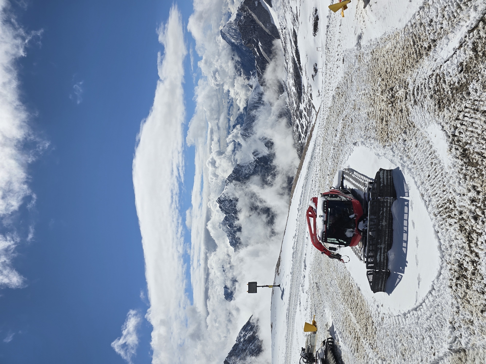 The image shows a snow groomer parked on a snowy mountain slope. In the background, majestic snow-covered mountains are visible, partially obscured by clouds, under a bright blue sky with scattered white clouds. The snow groomer is red and has a large track system. Other snow grooming equipment, including a yellow shovel, are also present on the slope. The terrain appears to be a ski resort, likely in the Zugspitze area near Garmisch-Partenkirchen, Germany, given the mountainous landscape. The scene is serene and untouched, with the snow groomer suggesting recent activity or preparation for use.