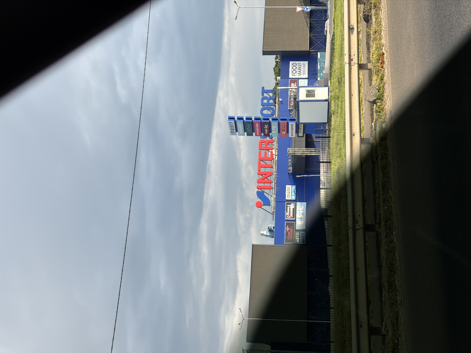 An exterior shot captures a large blue building with "INTERSPORT" emblazoned across the front in red and blue letters. To the left of the main sign is a tall, multi-colored vertical sign with various shop names listed, including "EPICENTER" at the top. To the right of the "INTERSPORT" sign, a smaller sign reads "FOOD MARKET FOOD COURT". The building appears to be a commercial or retail establishment, possibly a shopping center or mall, situated in a suburban or industrial area. A metal guardrail runs along the foreground, separating the road from a grassy area and the building. The sky is overcast with dark clouds, suggesting a possibility of rain. The overall lighting indicates it is daytime.