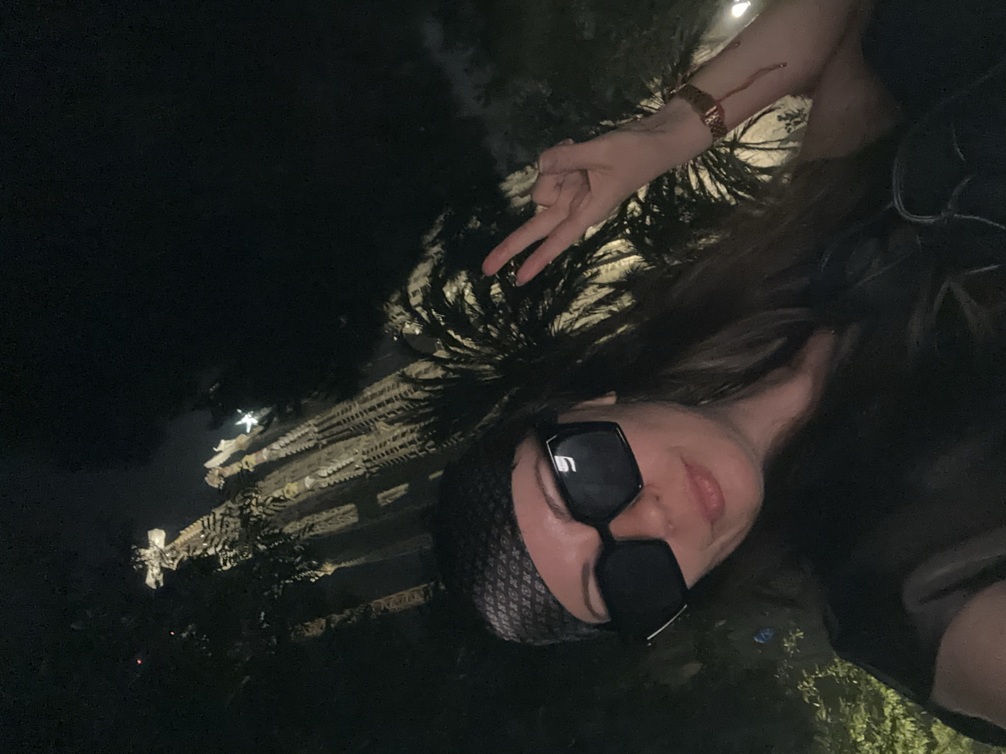 The image is a selfie taken at night. The subject is a person with long, dark hair wearing black, oversized sunglasses and a dark bandana with a repeating pattern. They are holding up two fingers in a peace sign. On their left wrist, they are wearing a rose gold watch. The background is dark with some ambient lighting, suggesting an outdoor setting at night. To the left of the subject, a large, ornate structure is visible, illuminated by lights. This structure appears to be a building or monument with intricate details and a tall, spire-like element. The foliage in the foreground is dark and appears to be palm trees, consistent with the location context of Barcelona, Spain. The lighting and atmosphere suggest a warm, clear night. The person's expression, with a slight smile and the peace sign, conveys a sense of joy or playfulness. The overall scene captures a moment of personal enjoyment while exploring a visually interesting location at night.