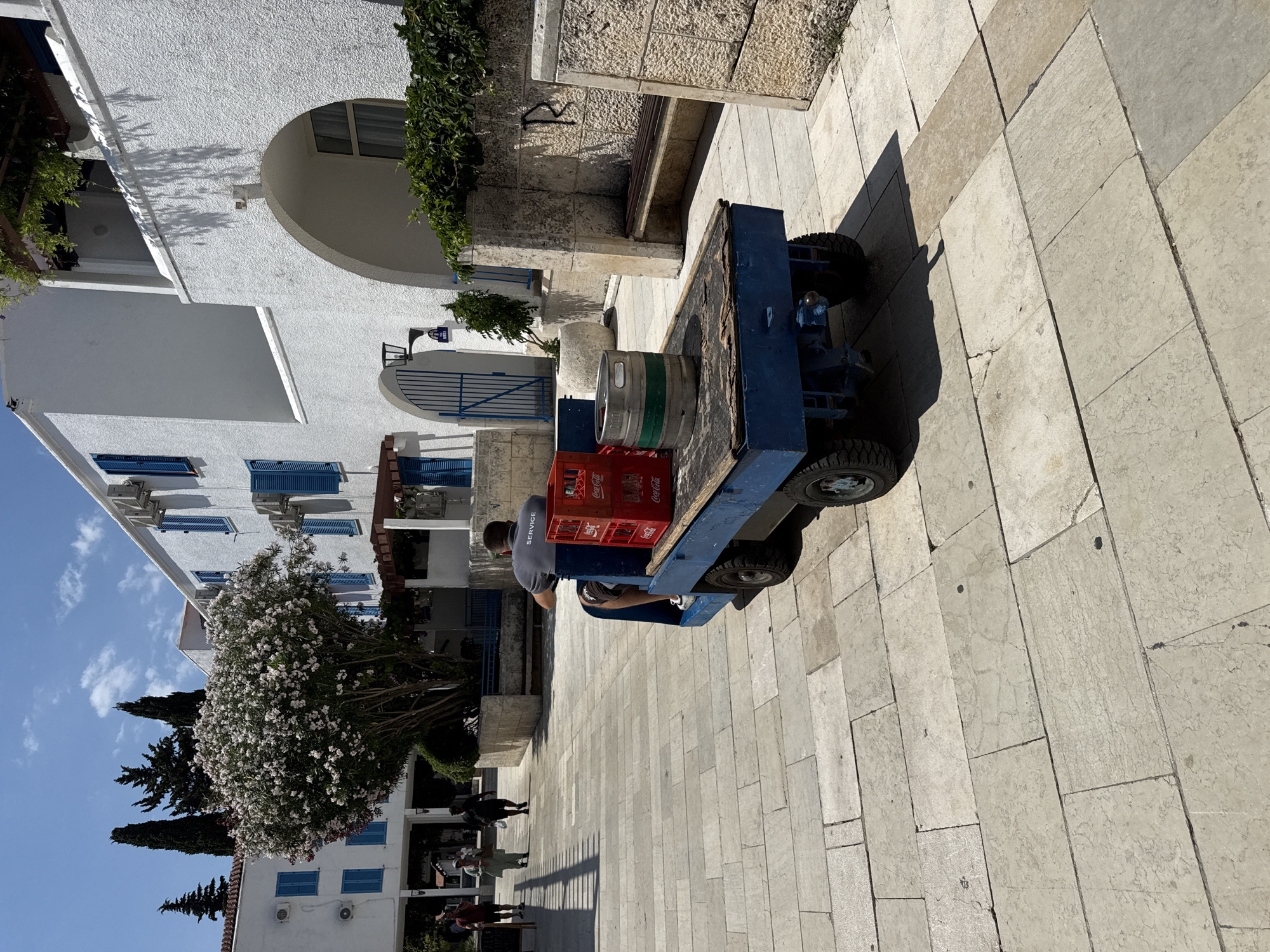 In Budva, Montenegro, a man wearing a grey t-shirt with the word "SERVICE" printed on the back is operating a small blue electric utility vehicle. The vehicle is transporting a red plastic crate filled with Coca-Cola bottles and a metal keg with green banding. The scene takes place on a paved pedestrian walkway made of light-colored stone slabs. To the right of the walkway, there is a stone wall with an arched opening and a wooden bench. Behind the walkway, a white building with blue shutters and air conditioning units is visible, along with a large tree in bloom. Several other pedestrians are walking in the background. The lighting suggests it is daytime.