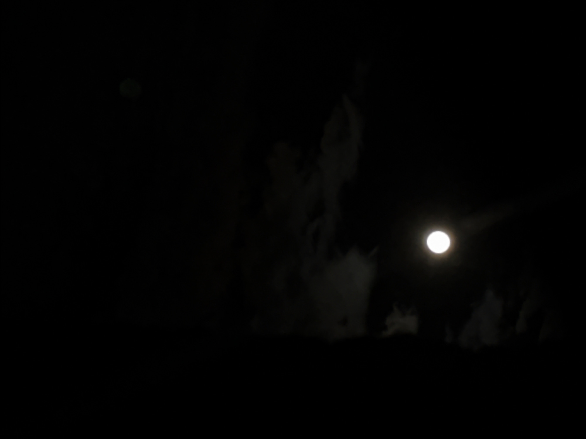The image displays a dark, nocturnal sky with a prominent, bright, circular light source, likely the moon, positioned in the right-center. This light is intense and white. Surrounding and partially shrouding the sky are faint, wispy, and amorphous cloud formations, appearing as light gray, translucent shapes. These atmospheric elements are particularly noticeable around the central and lower-right portions of the image. A much smaller, very faint, greenish circular light is also visible in the upper-left quadrant. The overall scene is characterized by deep darkness, emphasizing the illuminated moon and subtle cloud textures. No specific location cues, human figures, objects (other than celestial/atmospheric), or discernible activities are present. The time of day is clearly night, with cloudy weather conditions. There is no visible text.