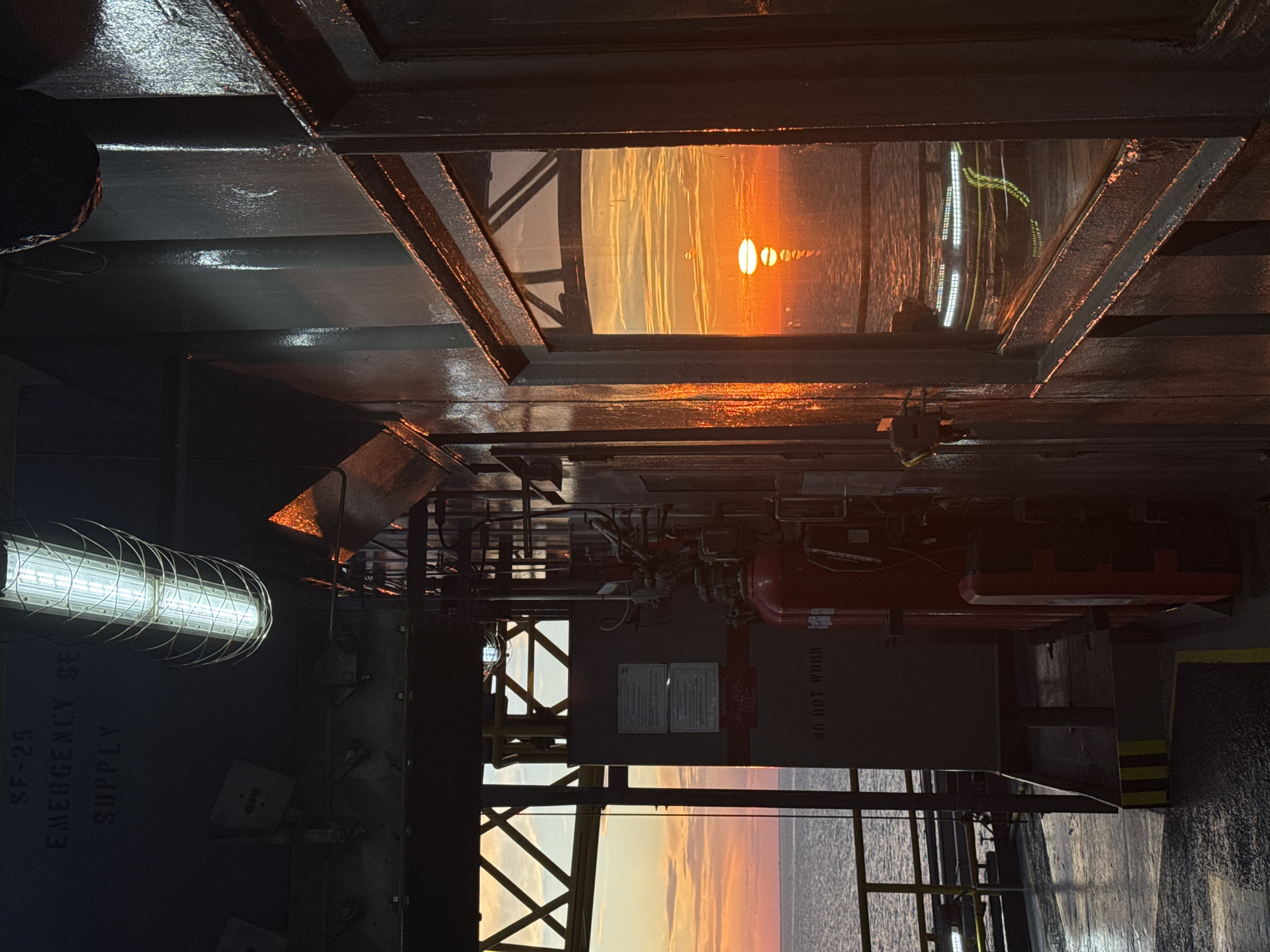 The image captures an industrial setting at sunset, possibly on an offshore platform. The sky is a vibrant orange and yellow, with the sun visible as a bright orb reflected in the water. In the foreground, there is a dark, weathered structure with a window reflecting the sunset. Inside the structure, industrial equipment is visible, including a red fire extinguisher and a control panel with warning labels. An overhead fluorescent light is also illuminated. The overall atmosphere is one of rugged industrialism juxtaposed with the natural beauty of the sunset. There are no people or animals present in the image. The location is specified as Unknown, Italy, but this appears to be a placeholder as the image depicts an offshore industrial environment, not a typical city scene in Italy.