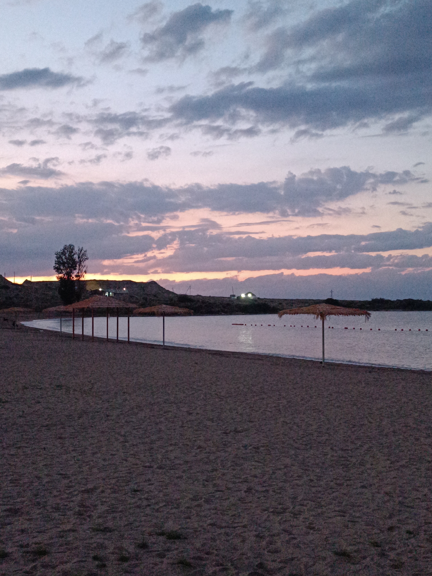 A desolate beach scene unfolds at dusk in Tong, Kyrgyzstan. The sand, appearing dark and somewhat uneven, stretches towards a tranquil body of water. Several thatched umbrellas, supported by poles, are sparsely dotted along the shoreline, suggesting an unoccupied resort or recreational area. Beyond the water, a low, hilly landscape is visible, with a single, dark tree standing prominently on the left side. Further in the distance, lights illuminate structures on the horizon, hinting at a settlement. The sky is a canvas of muted colors, with streaks of pale pink and orange from the setting sun intermingling with soft, gray clouds. The overall atmosphere is one of quiet solitude, typical of a beach at the end of the day.