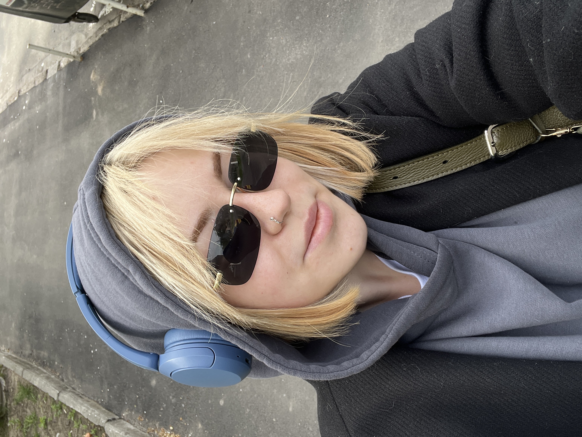 A close-up selfie captures a young person, likely female, outdoors in Odesa, Ukraine. The subject has short, bright blonde hair and is looking upwards towards the camera with a neutral expression, their eyes obscured by large, dark, aviator-style sunglasses. A small, silver hoop nose ring is visible on their left nostril.
They are wearing blue over-ear headphones, a grey hooded sweatshirt with the hood pulled up, and a dark, possibly black, outer jacket or coat. A dark olive green strap with a silver buckle, possibly belonging to a bag, is visible over their right shoulder.
The background shows grey asphalt or concrete pavement, indicating a street, sidewalk, or pathway. A concrete curb and patches of sparse green and brown vegetation are visible in the lower left corner. Some indistinct metallic structures can be seen in the upper left background. The lighting suggests it is daytime, possibly overcast or with diffuse sunlight, as shadows are soft and subtle. The overall impression is one of casual attire and a moment captured while listening to music or an audiobook outdoors. No discernible text is visible in the image.