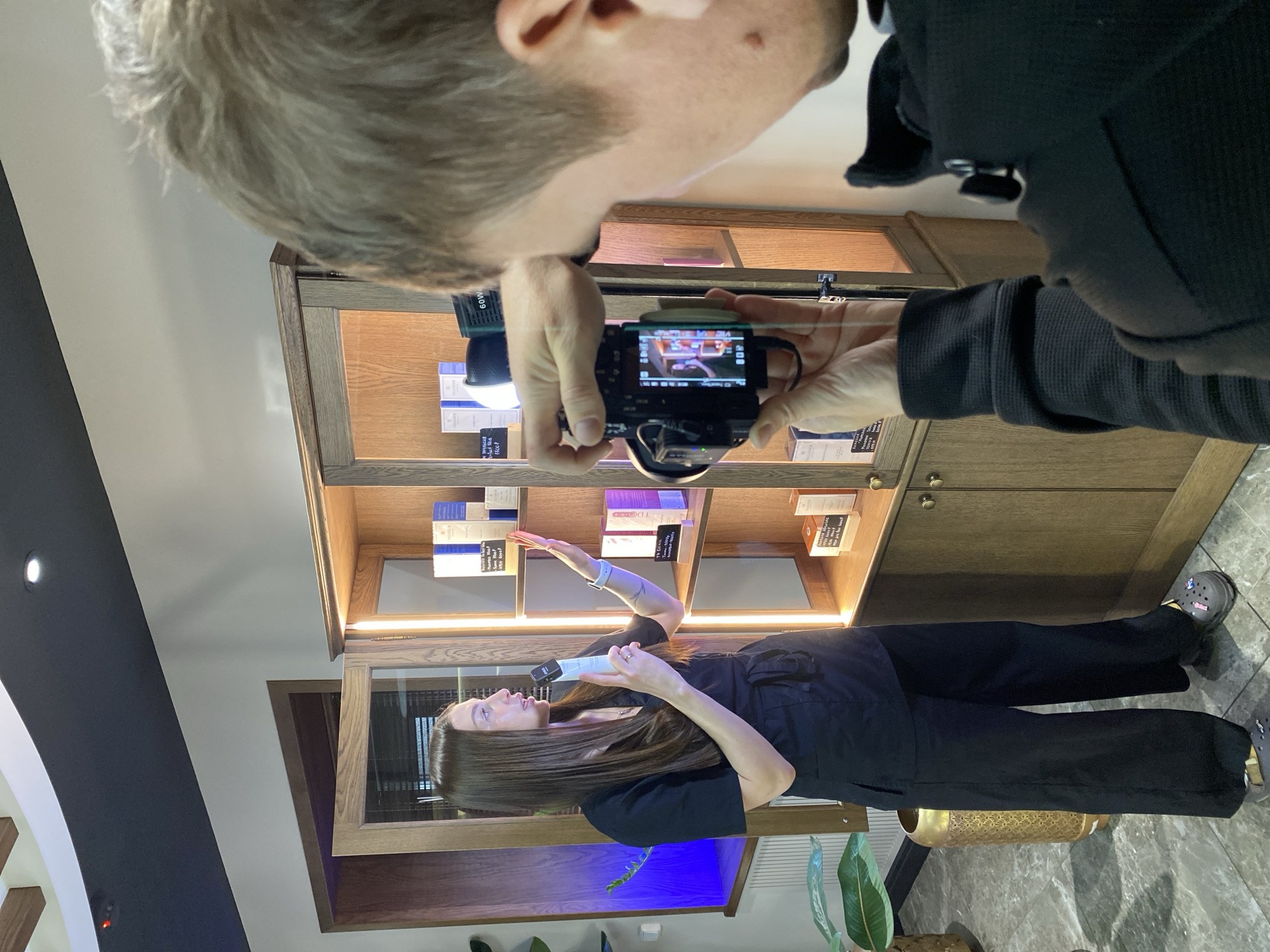 A medium shot of a person taking a photo of a woman holding a product in what appears to be a retail store.
People:
1. Person with fair skin, gray hair, wearing a black jacket. They are holding a camera.
2. A woman with long, brown hair. She is wearing a black outfit and holding a white object.
Objects:
1. A camera, being held by the person at the top of the image.
2. Display shelves with various products on them.
3. The white object being held by the woman.
4. A large green plant is visible in the bottom right corner.
Environment:
1. The scene appears to be inside a retail store, possibly a beauty or wellness shop.
2. The interior is well-lit, and the display shelves are made of wood.
Actions/Interactions:
1. The person is taking a photo of the woman.
2. The woman is presenting a product, possibly as part of a promotional activity.
Location: The location is not discernable as Votkinsk, Russia from the visible elements in this picture.