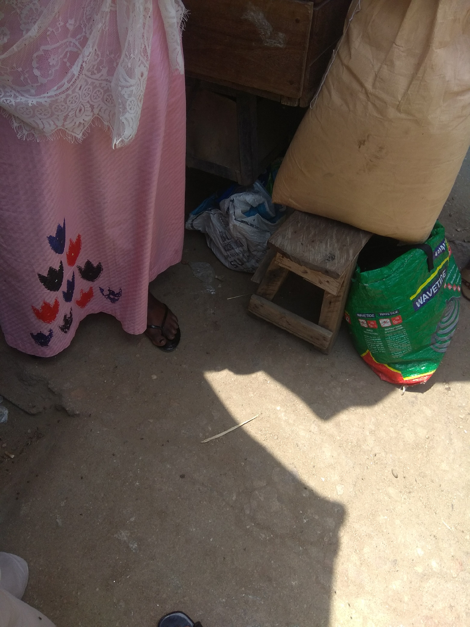 The image depicts a street scene in Jalingo, Nigeria. In the foreground, a person wearing a pink dress with lace trim and black flip-flops is partially visible, standing on a dusty ground. The dress features colorful floral patterns. To the right of the person, there is a large, light brown woven sack, a small wooden stool, and a green woven bag with text that appears to be "WAVETIDE" printed on it. Behind these objects, a wooden structure can be seen. The ground is made of concrete or compacted dirt, with some debris scattered around. Sunlight casts distinct shadows across the scene.