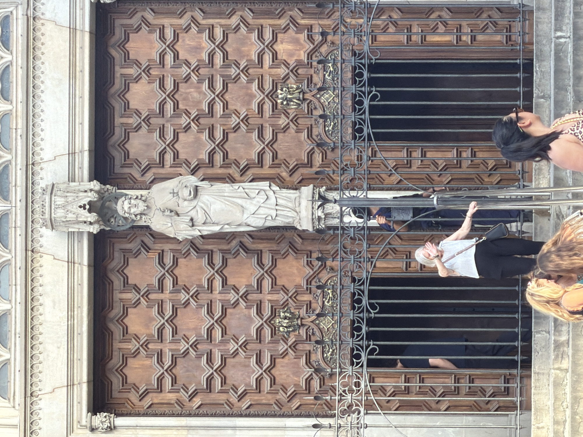 This is an outdoor, eye-level shot of a cathedral entrance in Barcelona, Spain. The main focus is a pair of large, ornate wooden doors featuring geometric patterns. A statue of a robed figure, possibly Jesus Christ, stands between the doors, holding a cross and orb. In front of the doors, a wrought-iron gate separates the entrance from a group of people on stone steps.
A woman in a white shirt and black pants stands behind the gate, shielding her eyes from the sun with her hand. To her left, a person in a dark blue shirt walks through the doorway. To the right of the gate, another person, possibly a guard, holds a pole. In the foreground, three women are visible on the steps, two of whom are looking towards the cathedral entrance.
The architecture suggests a historical building, consistent with many churches in Barcelona. The light suggests it is daytime, with the sun high in the sky.