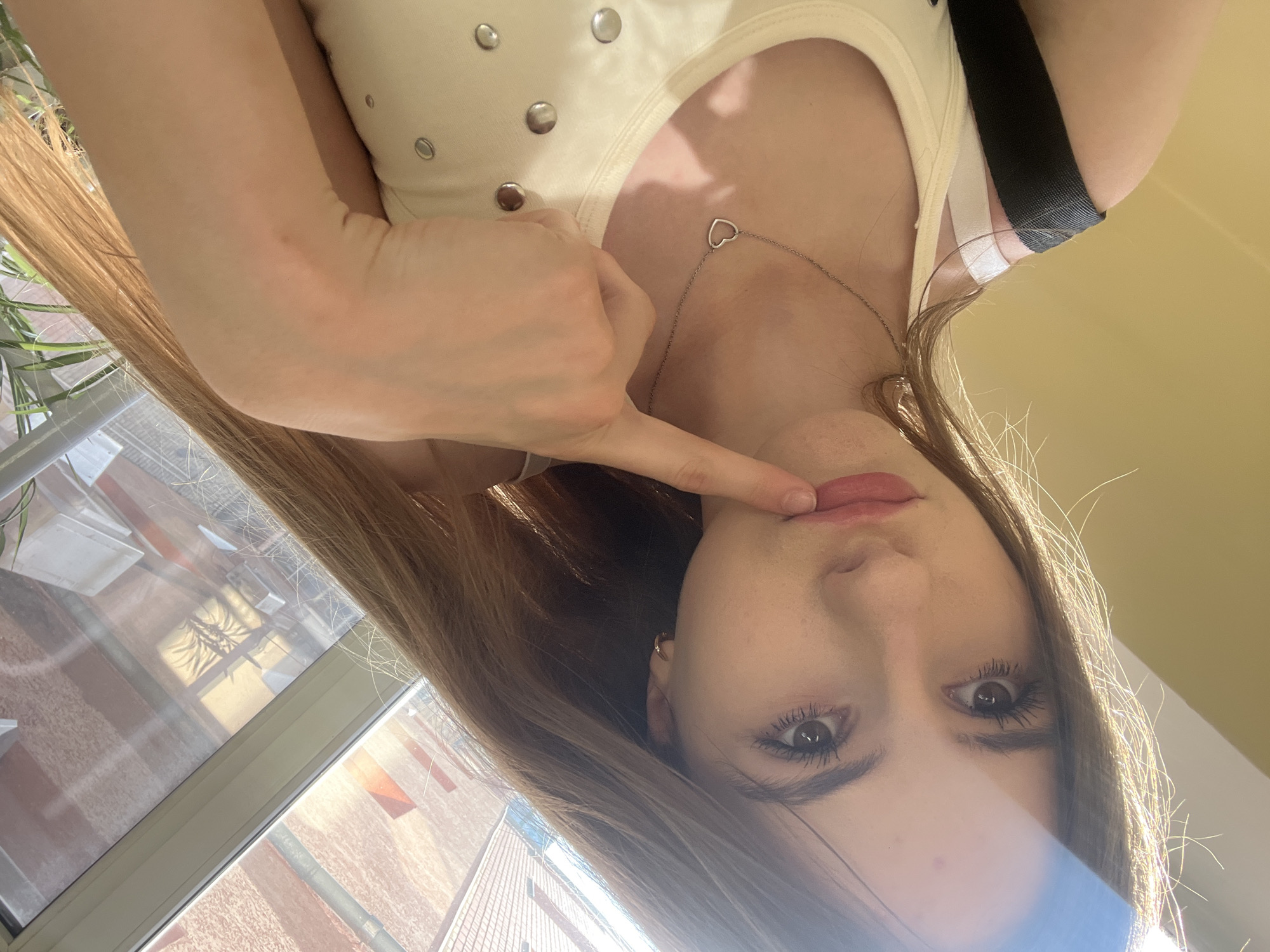 A young woman with long, light brown hair is taking a selfie, holding her phone at an unusual angle which causes the image to be rotated. She is wearing a cream-colored tank top with silver embellishments. A delicate silver necklace with a heart pendant rests on her collarbone. Her hand is raised, and her index finger is touching her lips, which are painted with pink lipstick. Her eyes are wide open and looking directly at the camera. The background suggests an indoor setting with large windows, through which a blurry view of buildings and possibly trees can be seen. The lighting indicates it's daytime. There is no visible text in the image. The overall impression is that of a playful or flirtatious selfie.