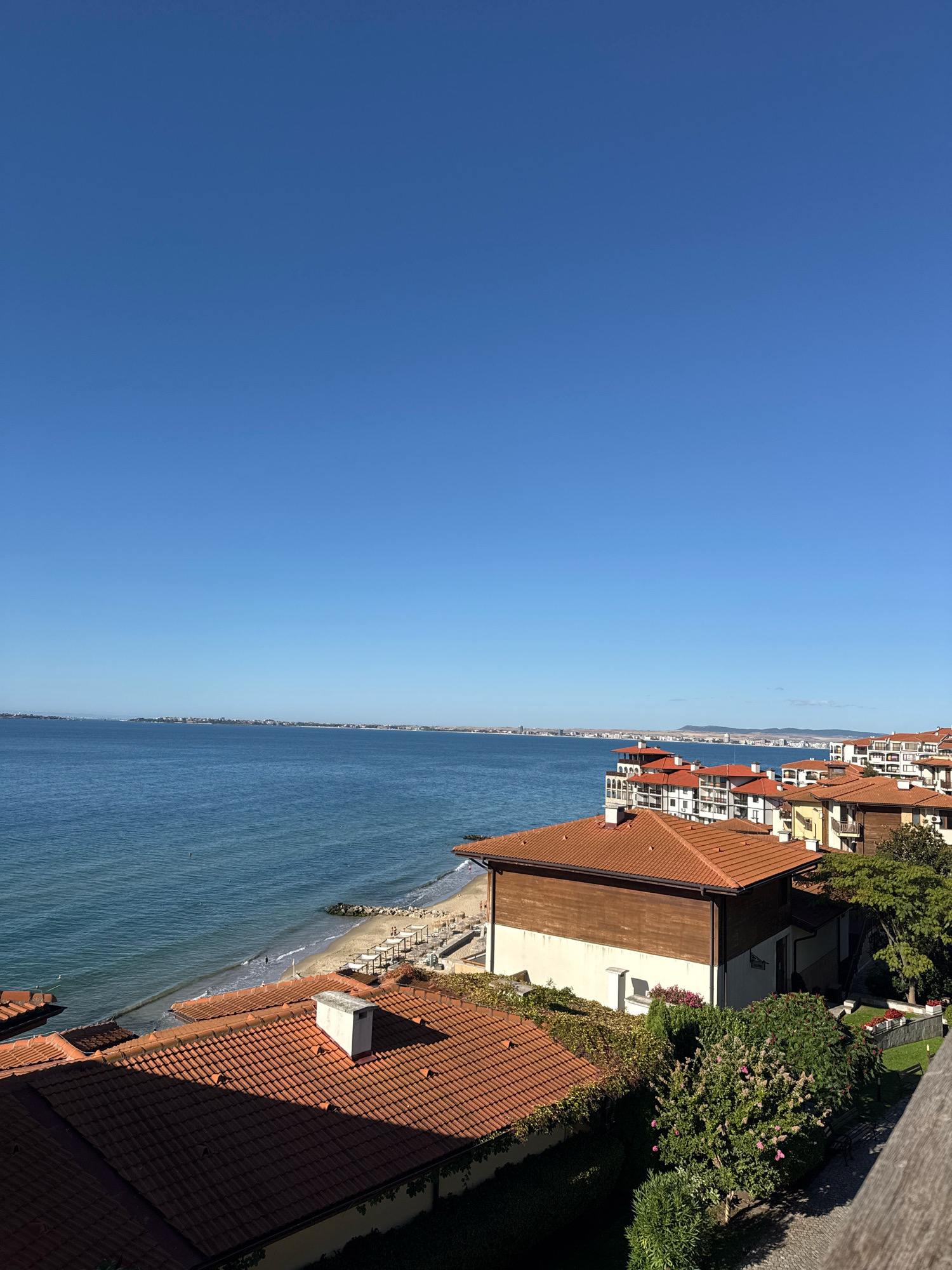 The image captures a scenic view of the Black Sea coast in Sveti Vlas, Bulgaria, on a clear, sunny day. In the foreground, several buildings with red-tiled roofs are visible, some covered with lush green ivy. Beyond these buildings lies a sandy beach with scattered lounge chairs and umbrellas, indicating a tourist season. The calm, blue sea stretches towards the horizon, where a distant cityscape with buildings and hills can be seen under a bright, cloudless blue sky. The overall impression is one of a tranquil coastal resort.