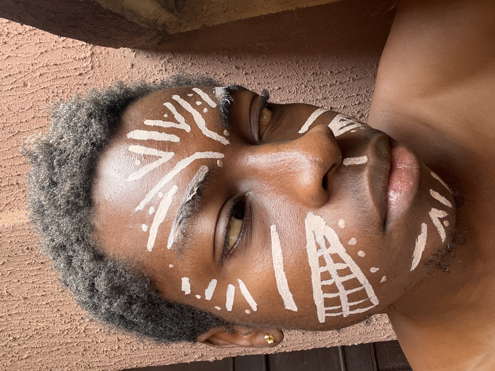 This close-up portrait features a person with dark skin and short, kinky dark hair, positioned against a textured, reddish-brown background. The individual's face is adorned with striking white body paint, composed of various geometric patterns, lines, and dots. Notable patterns include radiating lines on the forehead, a triangular grid-like design on the right cheek, and scattered dashes and dots across the face, including the bridge of the nose and around the eyes.
The person's eyes are partially open, looking subtly to the right, conveying a calm and contemplative expression. A small, gold-colored stud earring is visible in the left earlobe. The background consists of a rough, textured surface, possibly a stucco or concrete wall, showing variations in reddish-brown hues, along with darker, shadowed areas that suggest depth or other structural elements. The natural lighting indicates a daytime setting.
The presence of the detailed face paint suggests a cultural ritual, an artistic performance, or a creative photoshoot. Given the provided context, this scene is set in Nkpor, Nigeria, highlighting a potential traditional or contemporary artistic practice. No visible text is present in the image.