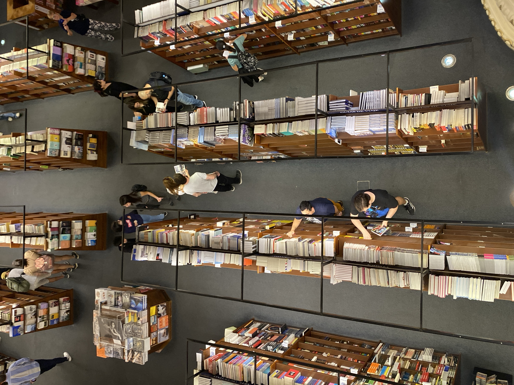 The image depicts the interior of El Ateneo Grand Splendid bookstore, located in Buenos Aires, Argentina, viewed from an elevated perspective. The scene features a multitude of dark wood bookshelves, densely packed with books, arranged in long, parallel rows that define aisles on a dark grey carpeted floor. Several square, central display units also hold books. Approximately twenty-five individuals, primarily adults, are present across the visible areas. People are engaged in various activities, including browsing books on shelves and display units, walking through aisles, and interacting with the merchandise. Specific actions include individuals reaching for books, holding open books, and conversing near shelves. Several individuals carry backpacks. Circular, recessed floor lights are visible in the carpet. Metal framework structures are present, delineating viewpoints and supporting the shelving.
