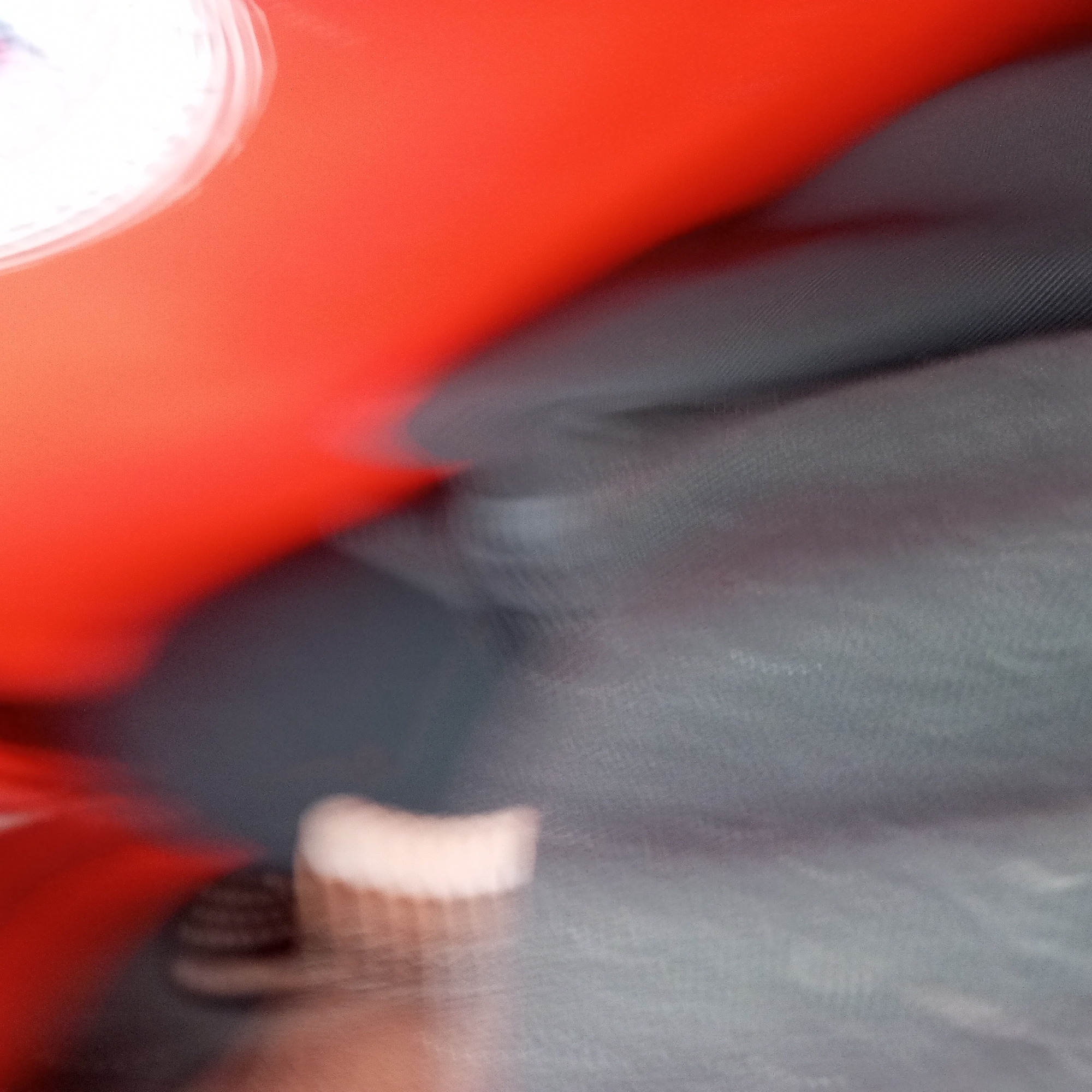This is an abstract, heavily blurred photograph dominated by vibrant red-orange and dark grey hues. A bright, circular white light source is visible in the top left, diffusing into the red-orange area. The upper portion of the image is characterized by a smooth, bright red-orange surface that extends diagonally across the frame.
The lower and central sections are occupied by a dark grey material. In some areas, particularly noticeable in the cropped views, this grey material displays a distinct linear or woven texture, suggesting it is a fabric like upholstery, a carpet, or part of a bag. The blurring across the entire image indicates significant motion or camera shake during capture, creating streaky patterns and obscuring fine details.
Towards the bottom center, a small, light beige or off-white object is partially visible. It appears to have vertical ridges or a textured surface, possibly belonging to a shoe, a brush, or another small item, but its identity is unclear due to the blur. No identifiable people, faces, or specific activities are discernible. The scene appears to be an interior space, potentially a vehicle or a room, given the combination of surfaces and the internal light source. There are no visible location cues or text, and the time of day or weather cannot be determined from the image. The photograph primarily functions as an abstract study of color, light, and texture.