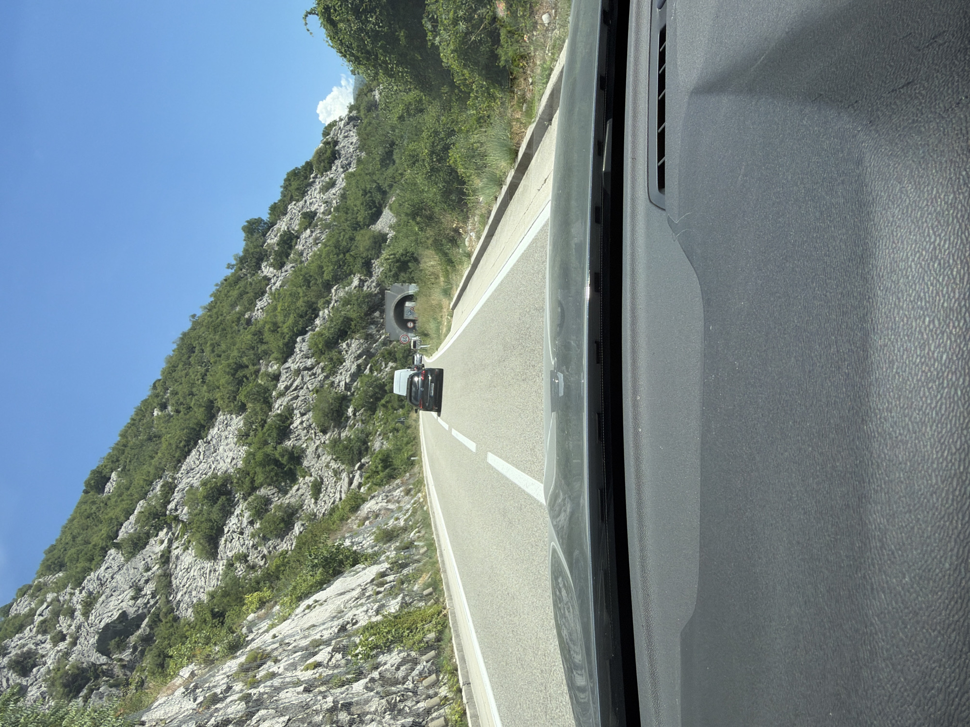 The image captures a view from inside a vehicle looking out at a road leading towards a tunnel. The road is paved and has white lane markings. On the left side of the road, a rocky, green-covered mountain slope rises steeply. On the right side, there is also dense green vegetation and a rocky incline. Several cars are visible on the road, including a dark-colored car in the foreground and a white van further ahead. A tunnel entrance is visible in the distance, with a speed limit sign of 60. The sky is clear and blue with a few scattered white clouds. The dashboard of the vehicle is visible in the lower part of the image, partially obscuring the view. The scene is consistent with a road passing through a mountainous area in Lipci, Montenegro.